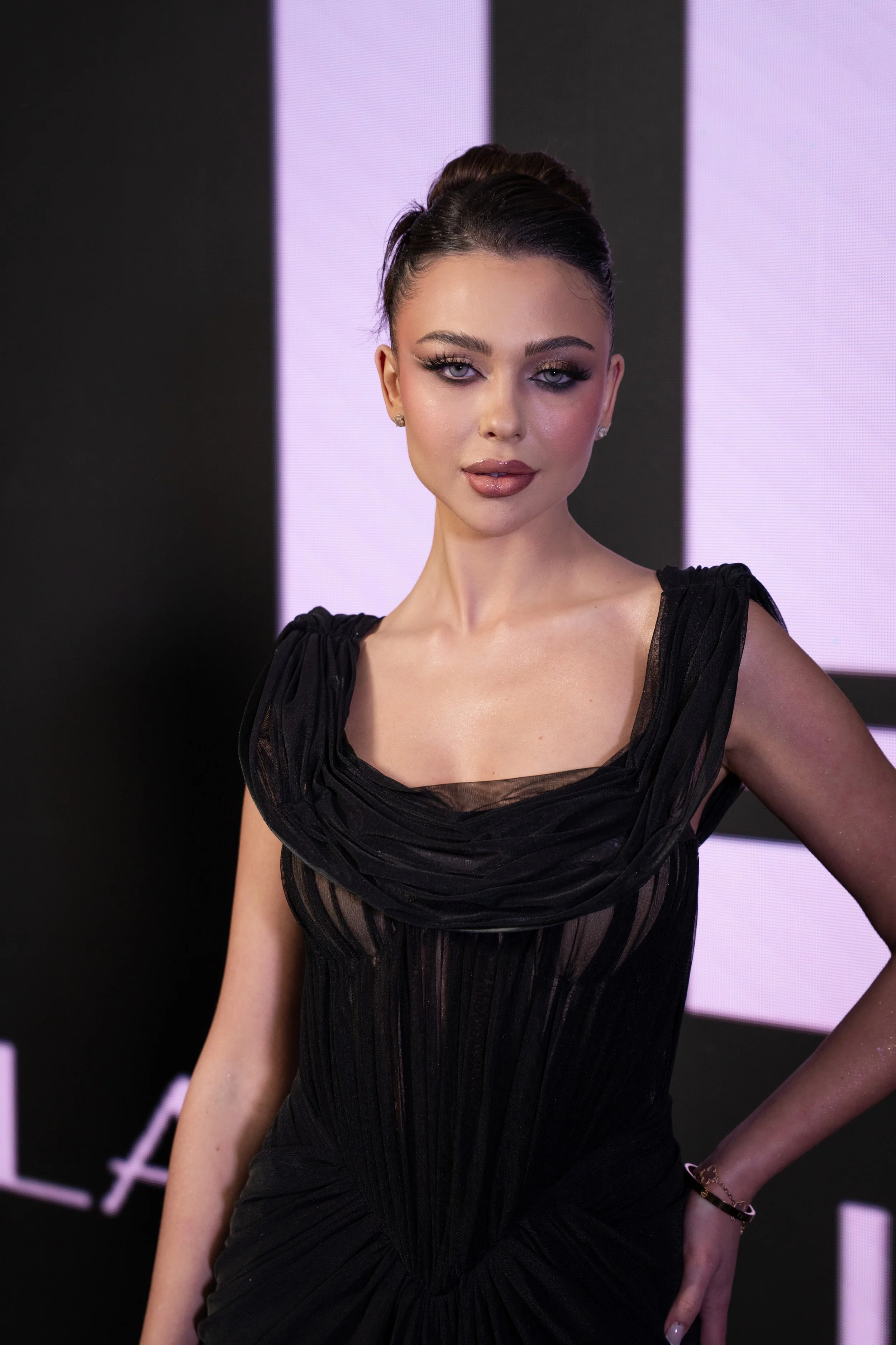 A woman with dark hair styled in an elegant updo, wearing a black dress with sheer and ruched details, and posing in front of a black and pink background.