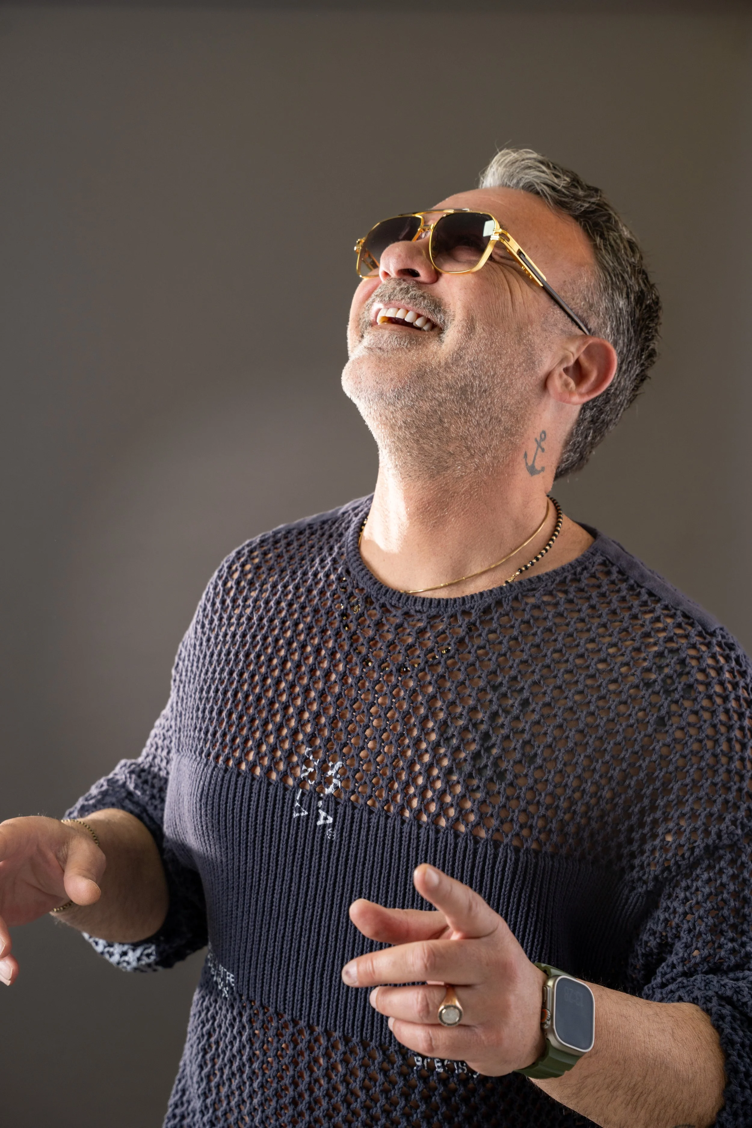 A smiling man with sunglasses, a grey beard, and a tattoo on his neck, wearing a navy blue mesh sweater, jewelry, and a smartwatch, is laughing and gesturing with his hands.
