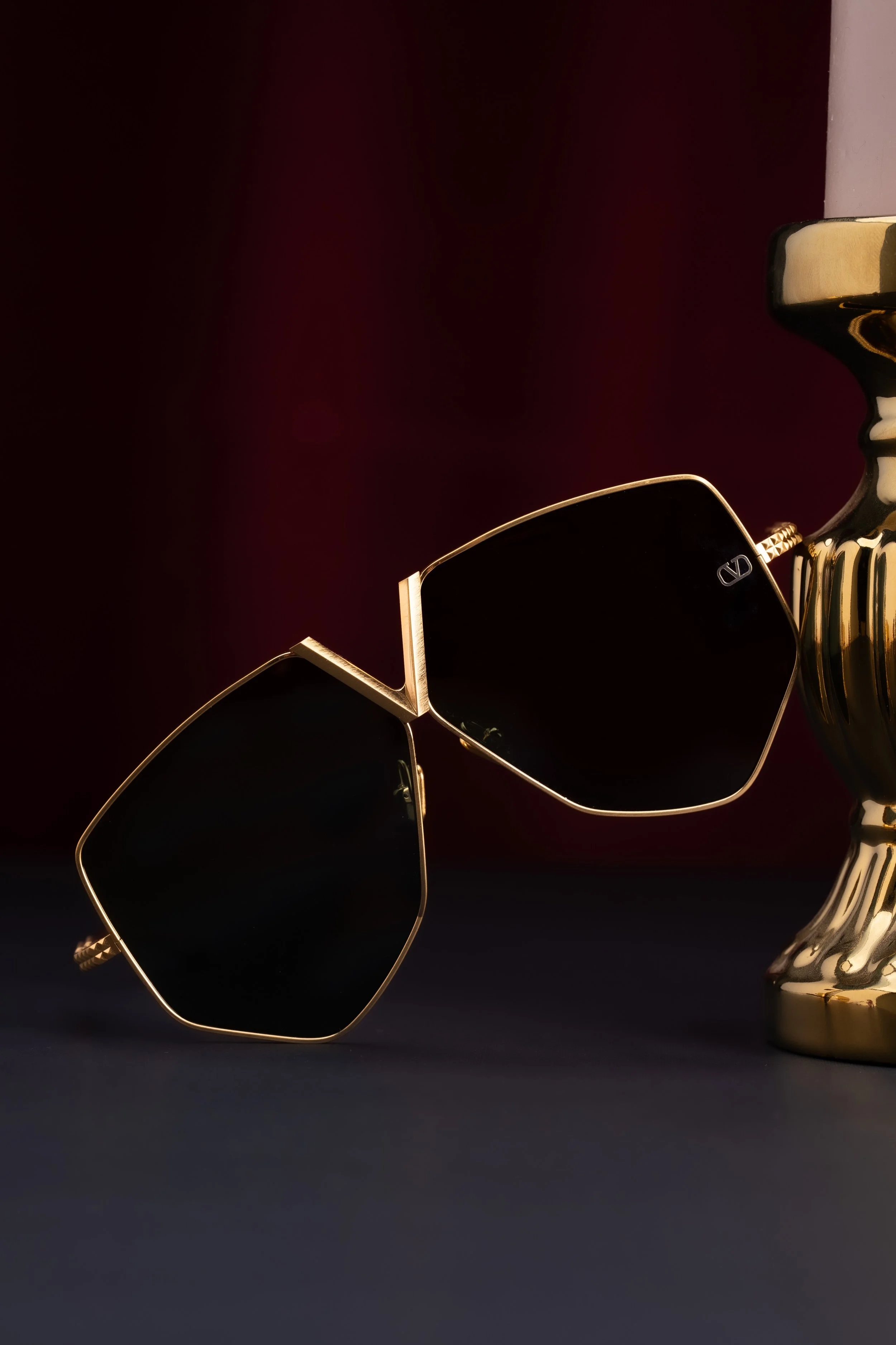 A pair of black sunglasses with gold frames resting on a dark surface next to a gold-colored candlestick.