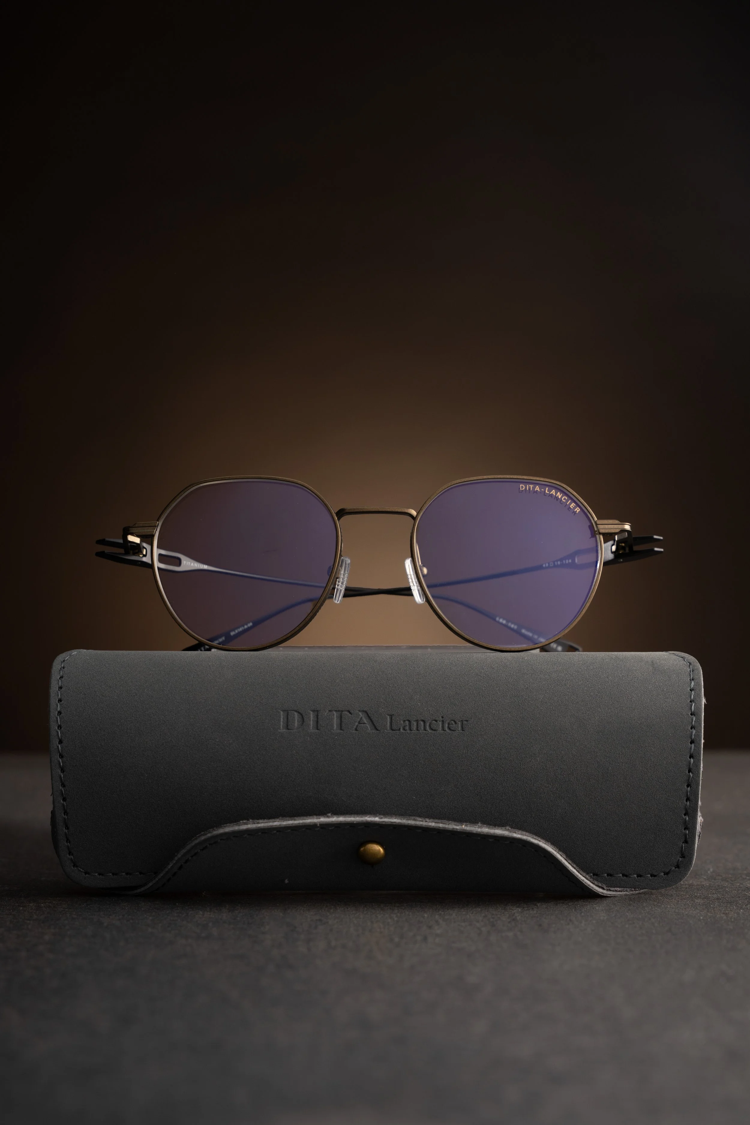 A pair of round sunglasses with purple lenses resting on a gray case with the brand name 'DITA Lancier' embossed on it.