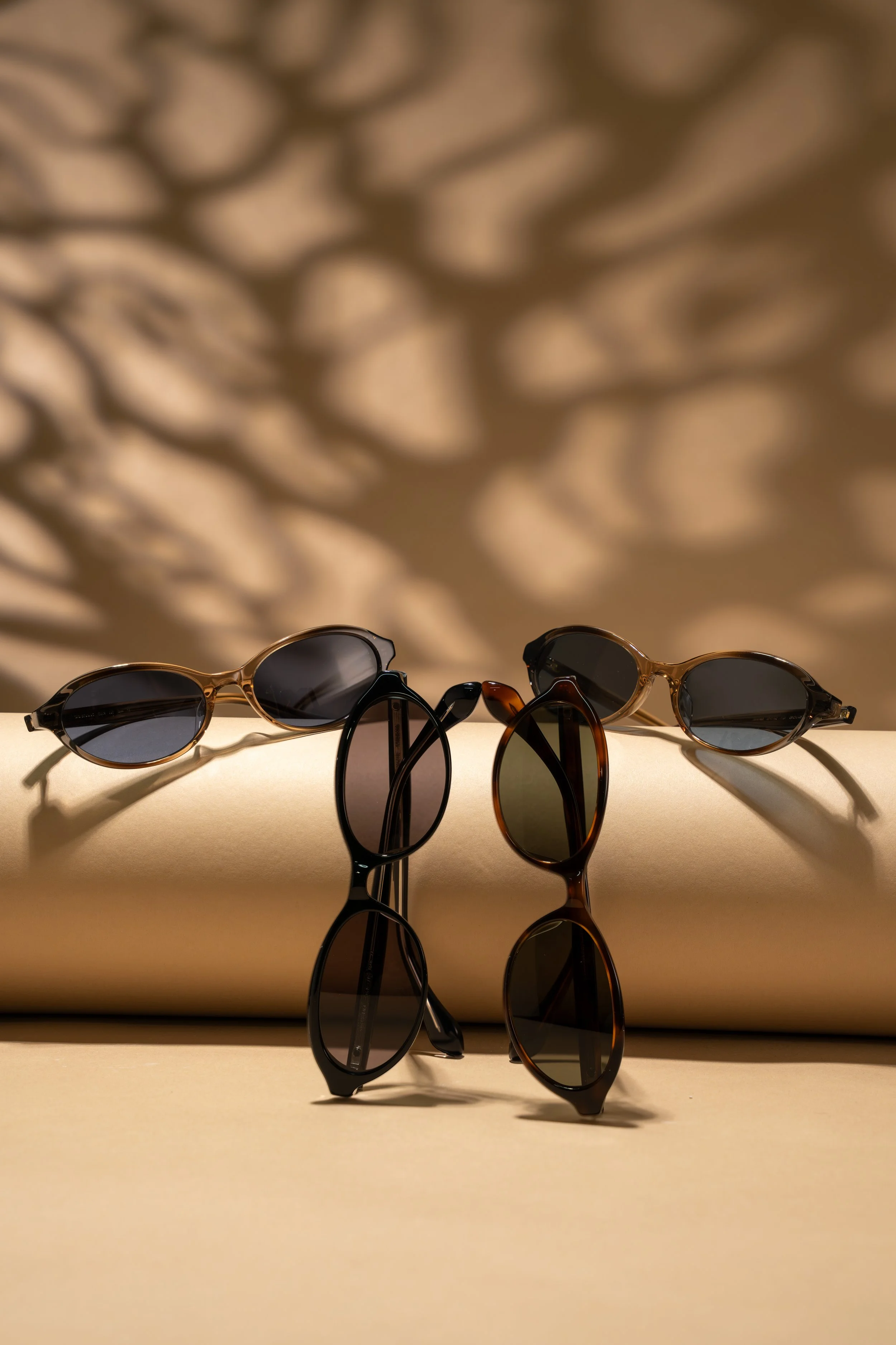 Three pairs of sunglasses with dark lenses and different frame styles are arranged on a beige surface with a mirror reflection, behind a background with intricate shadows of leaves cast on it.