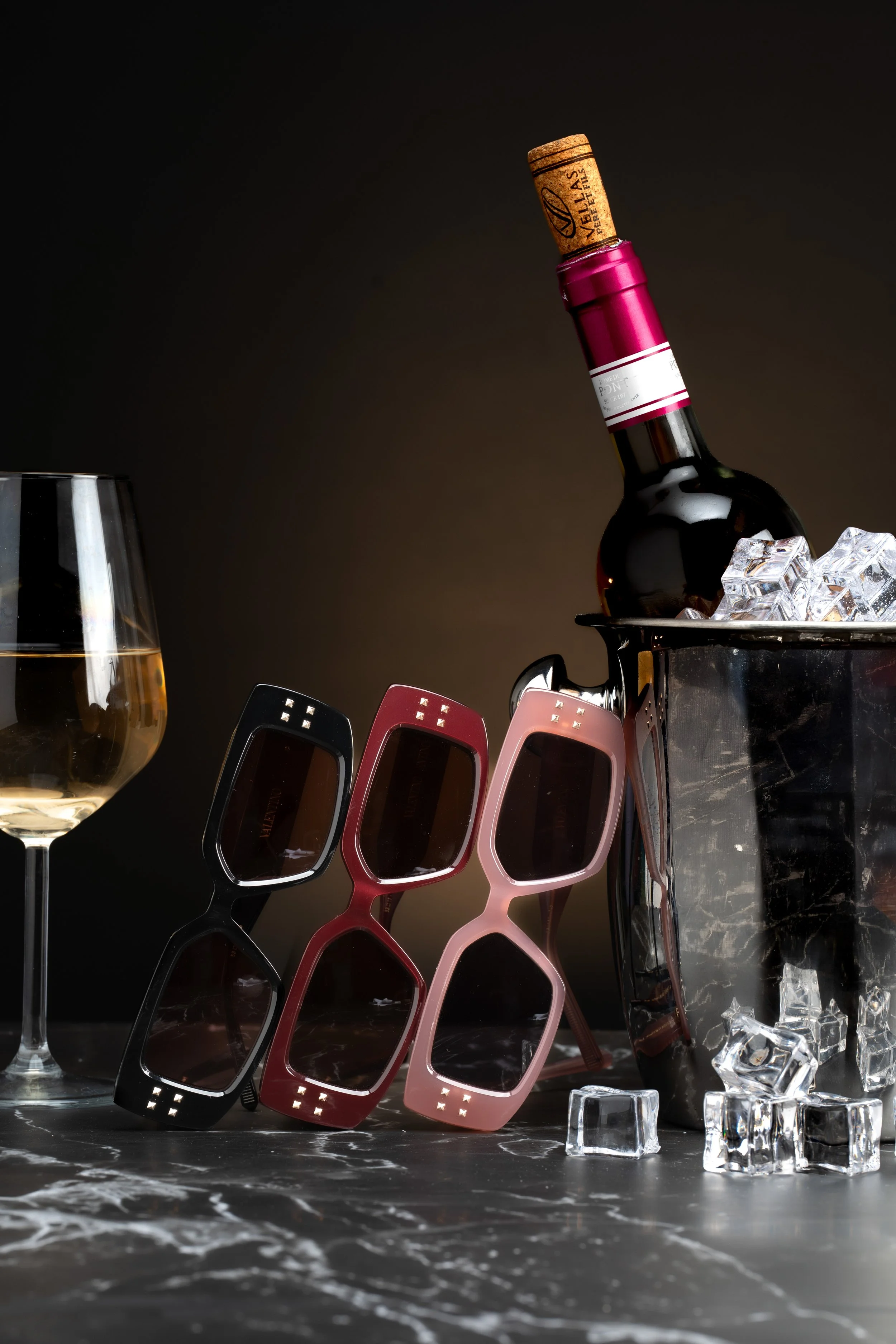 A glass of white wine, three pairs of sunglasses in black, red, and pink, a wine bottle in an ice bucket with ice cubes, and a dark background with a marble surface.