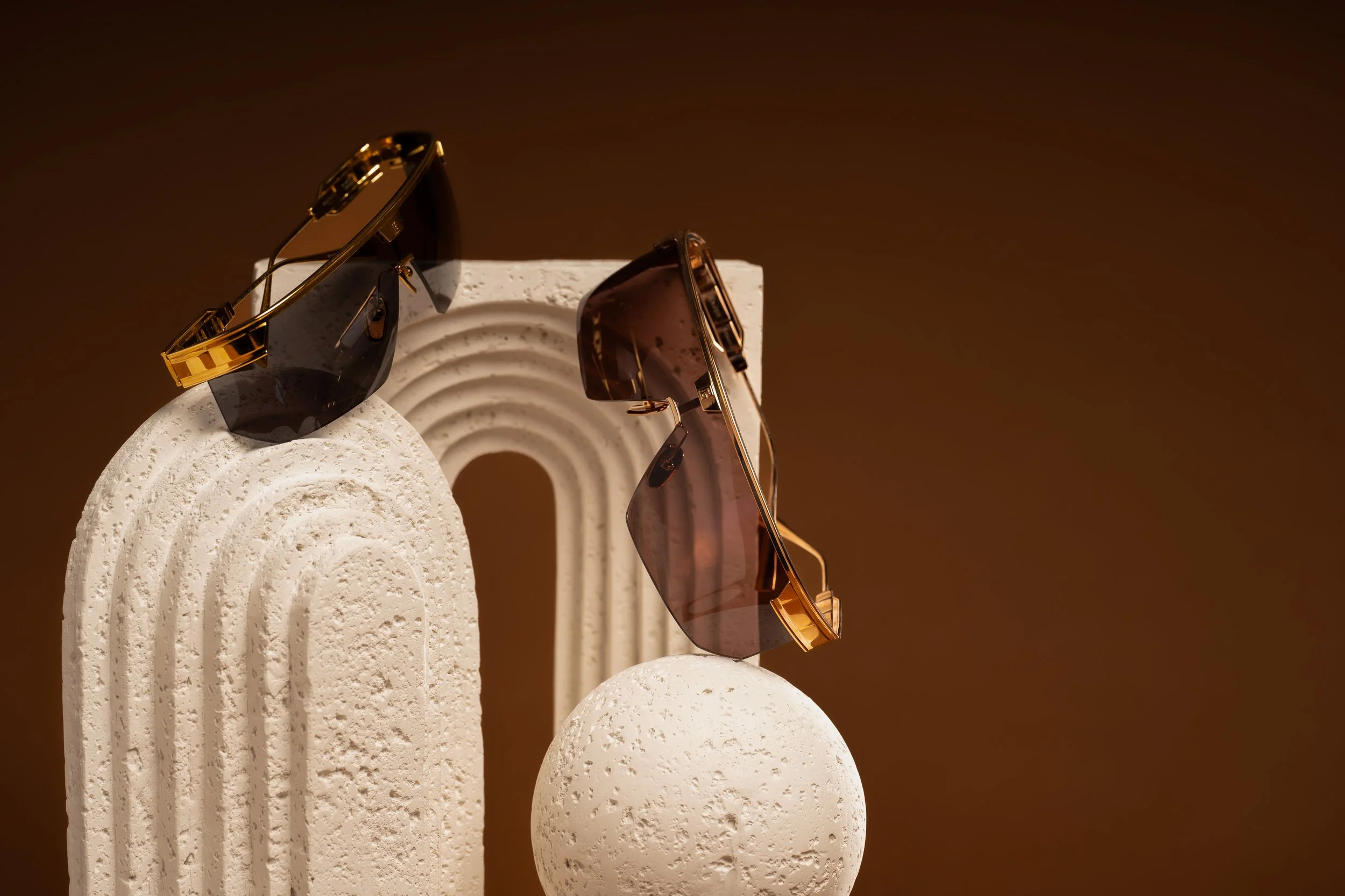 Two pairs of sunglasses with dark lenses and gold frames resting on textured white geometric objects, including a tall arched shape and a smaller sphere, against a brown background.