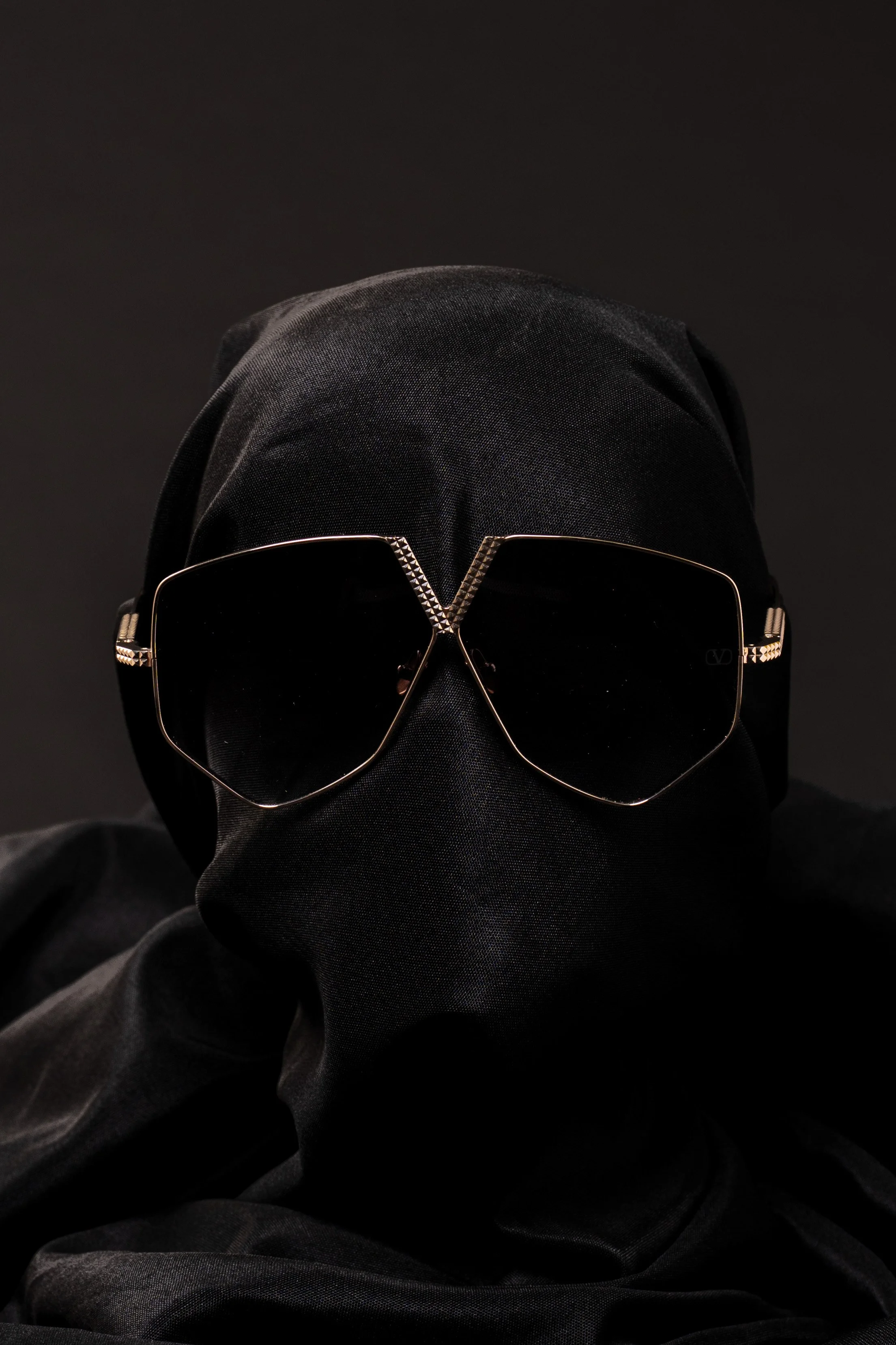 A black mannequin head wearing black sunglasses and a black wrap or hood, set against a dark background.