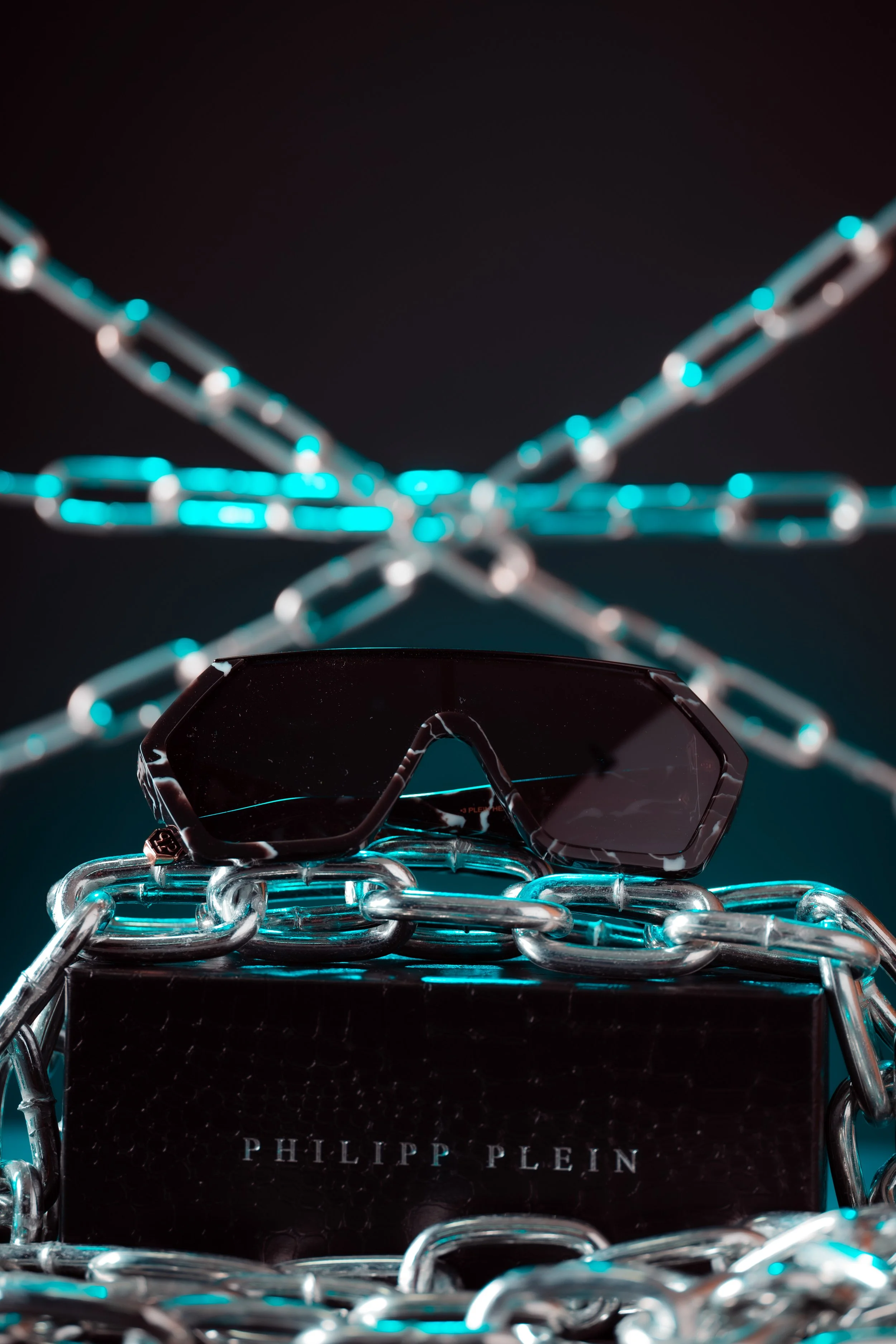 A pair of Philippe Plein sunglasses resting on a black box, surrounded by silver chains with chains in the background.