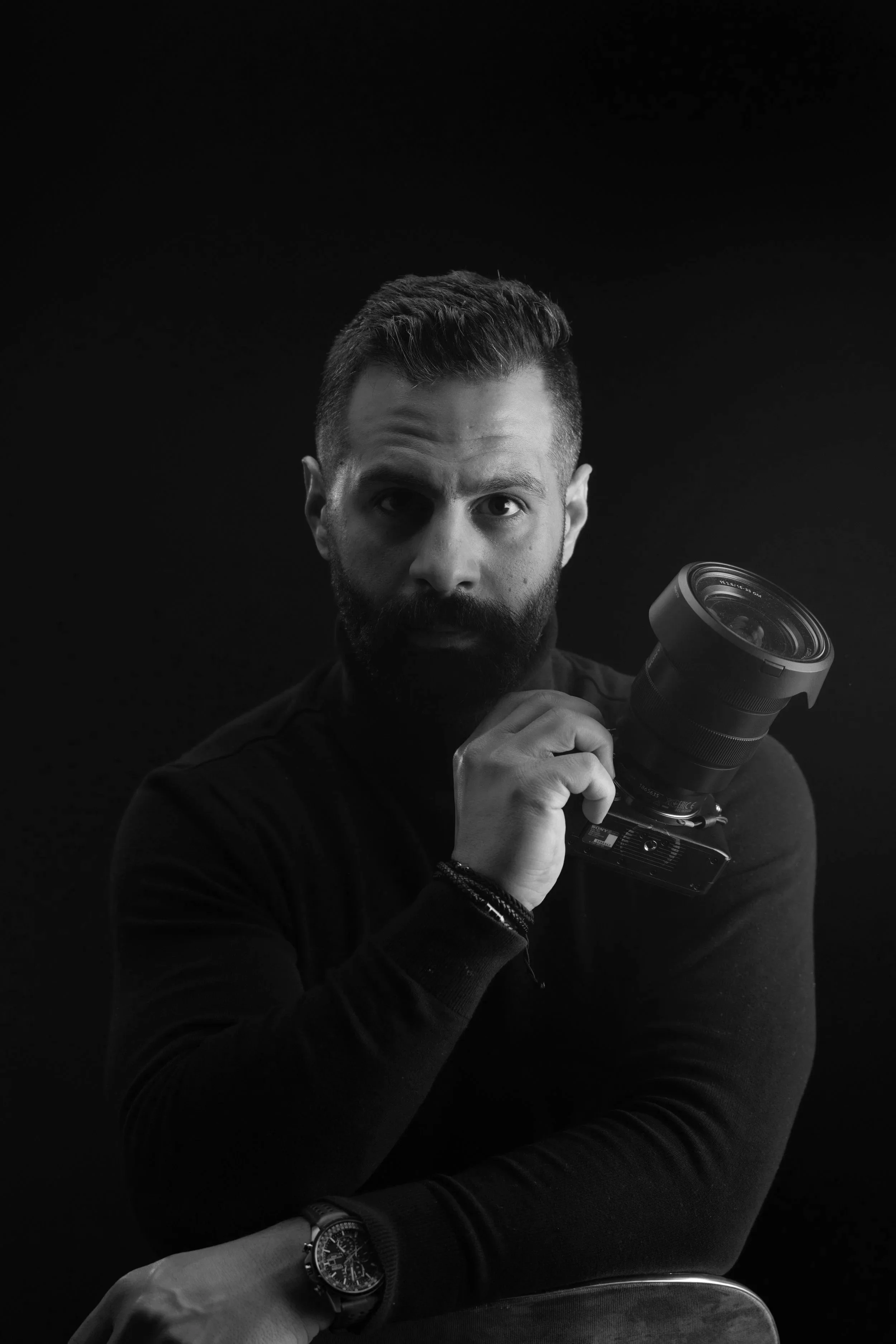 Black and white photo of a man with a beard and short hair holding a professional camera with a large lens, against a dark background.