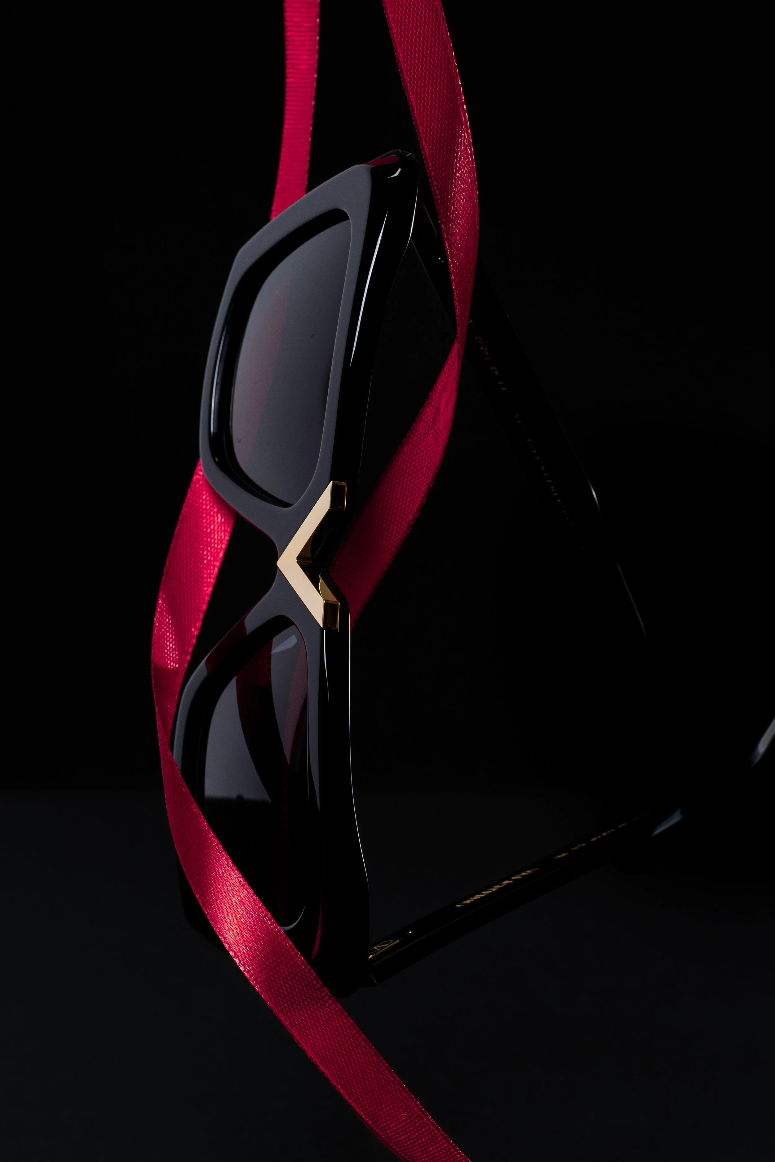 Black sunglasses with gold accents hanging on a red ribbon against a black background.