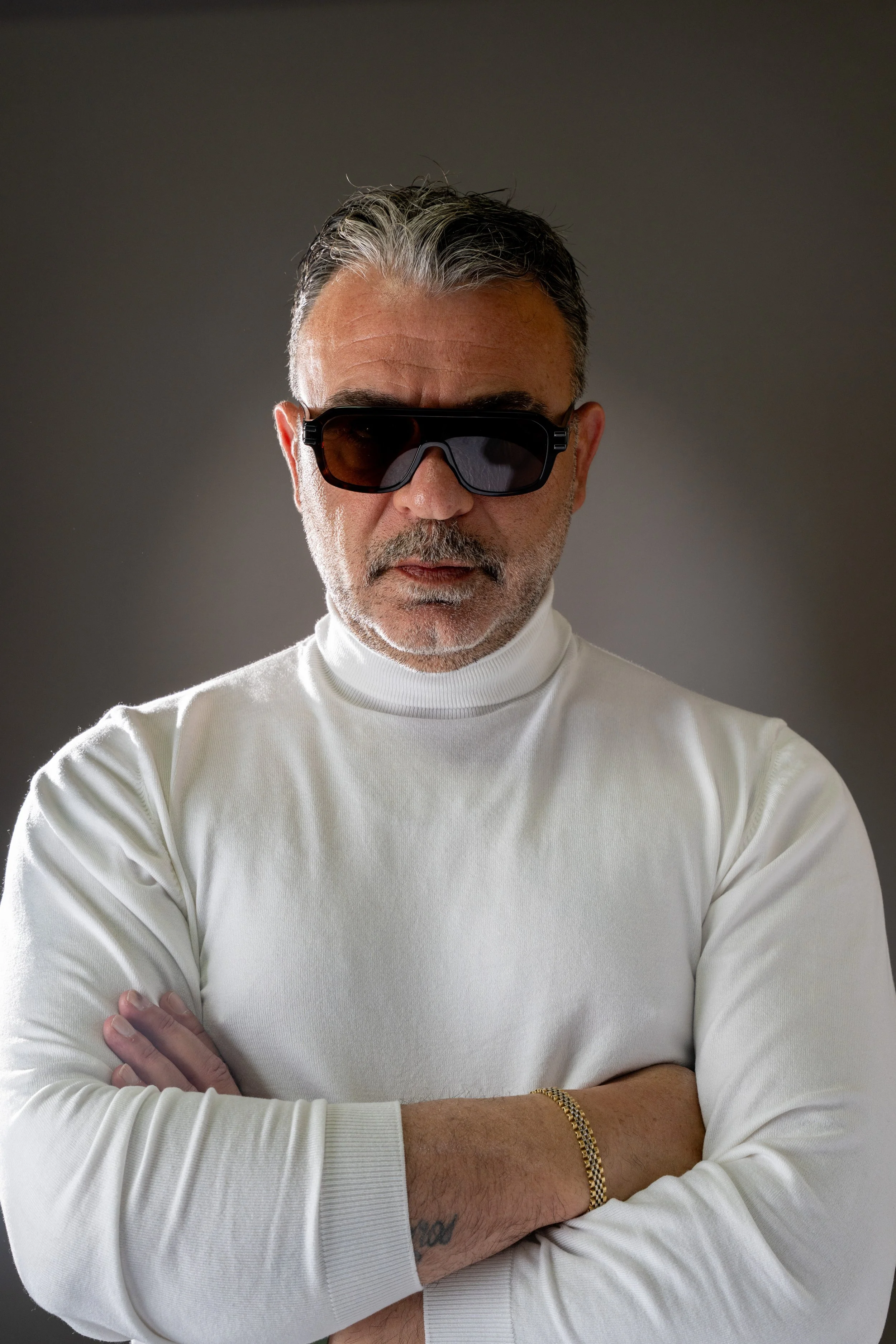 A man with gray hair and a beard wearing black sunglasses and a white turtleneck, crossing his arms and looking at the camera with a serious expression, against a dark background.