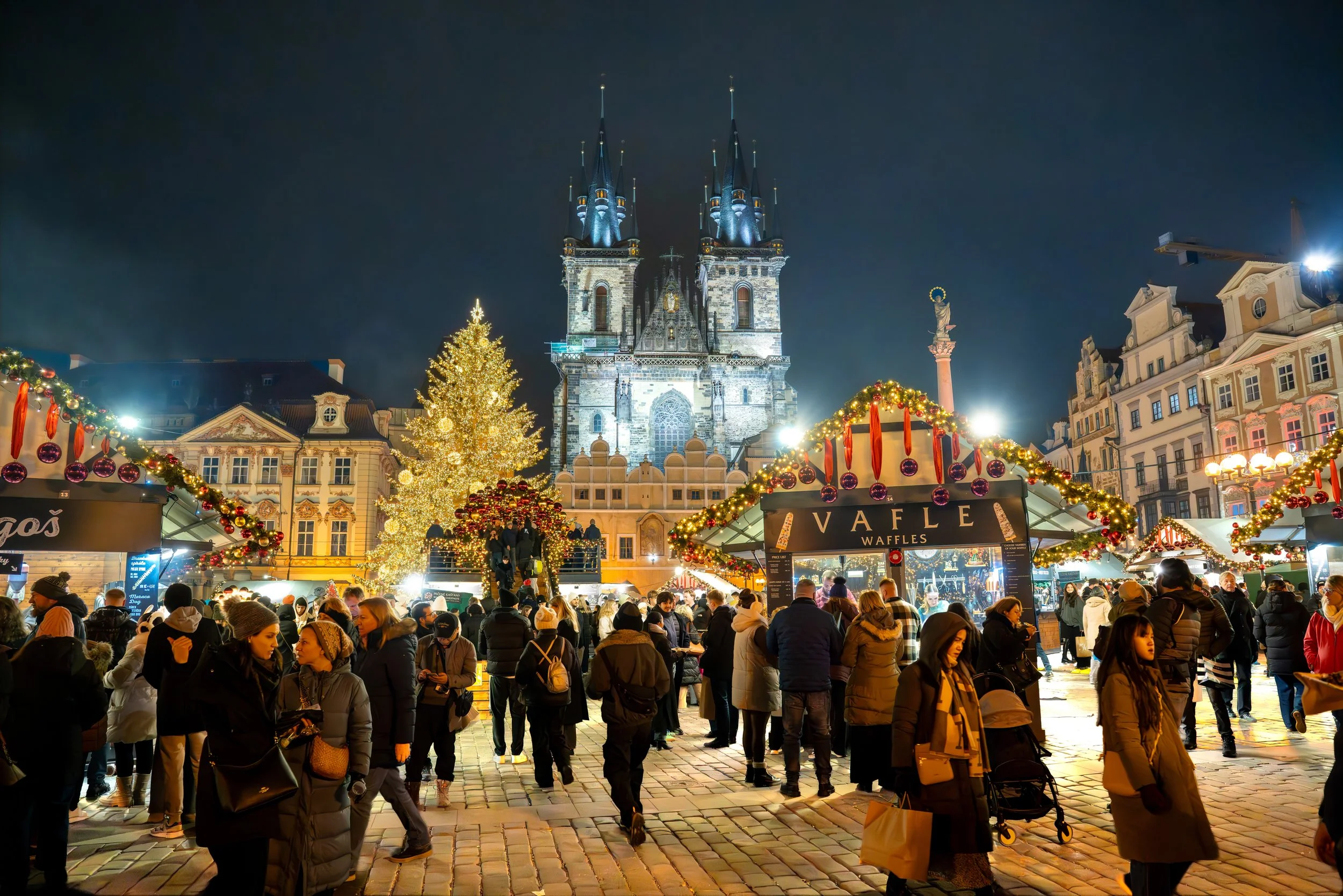 Nighttime Christmas market in a European square with a large decorated Christmas tree, festive stalls, and a crowd of people, with a historic church in the background.
