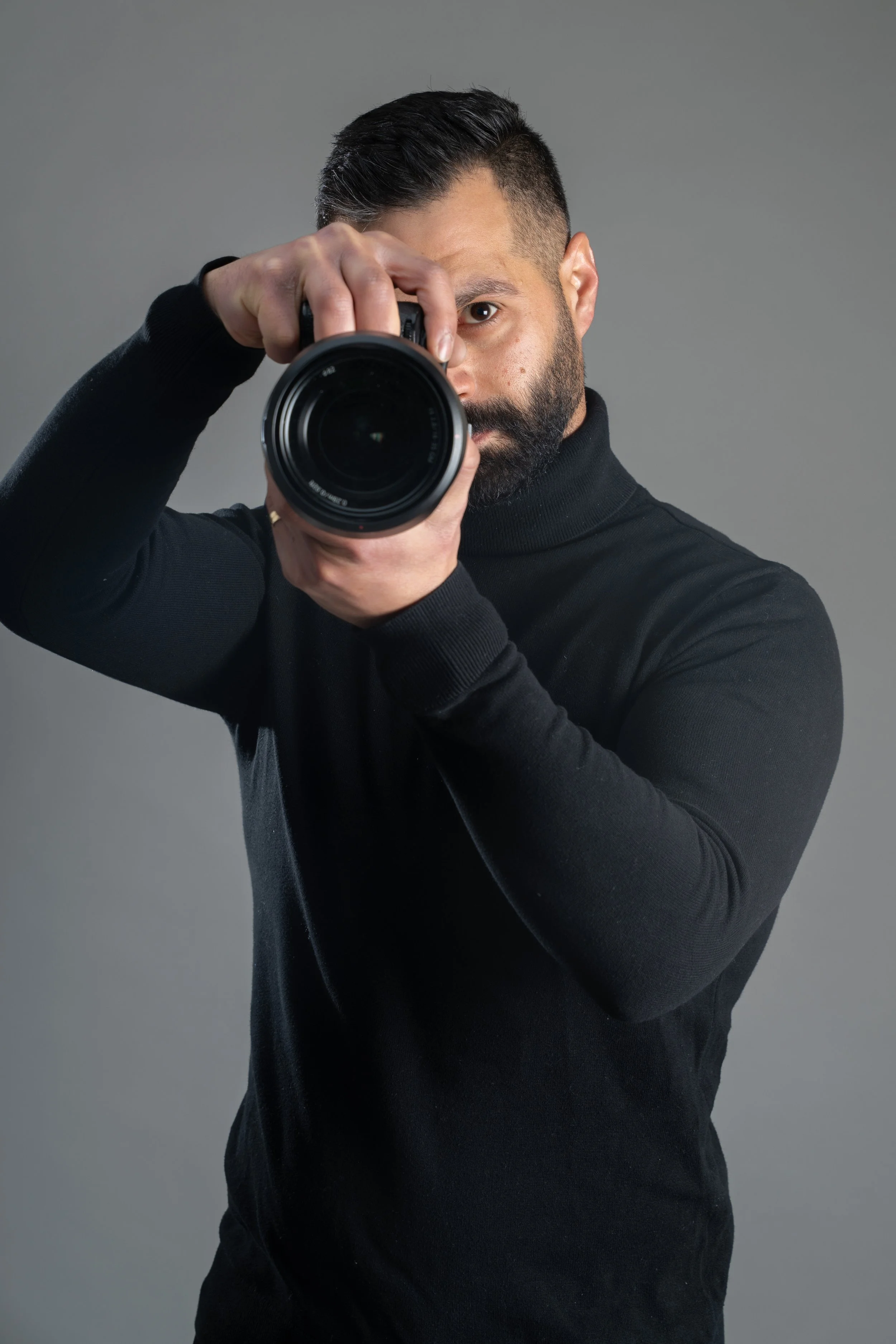 Man with dark hair and beard taking a photo with a camera, wearing a black turtleneck, against a gray background.