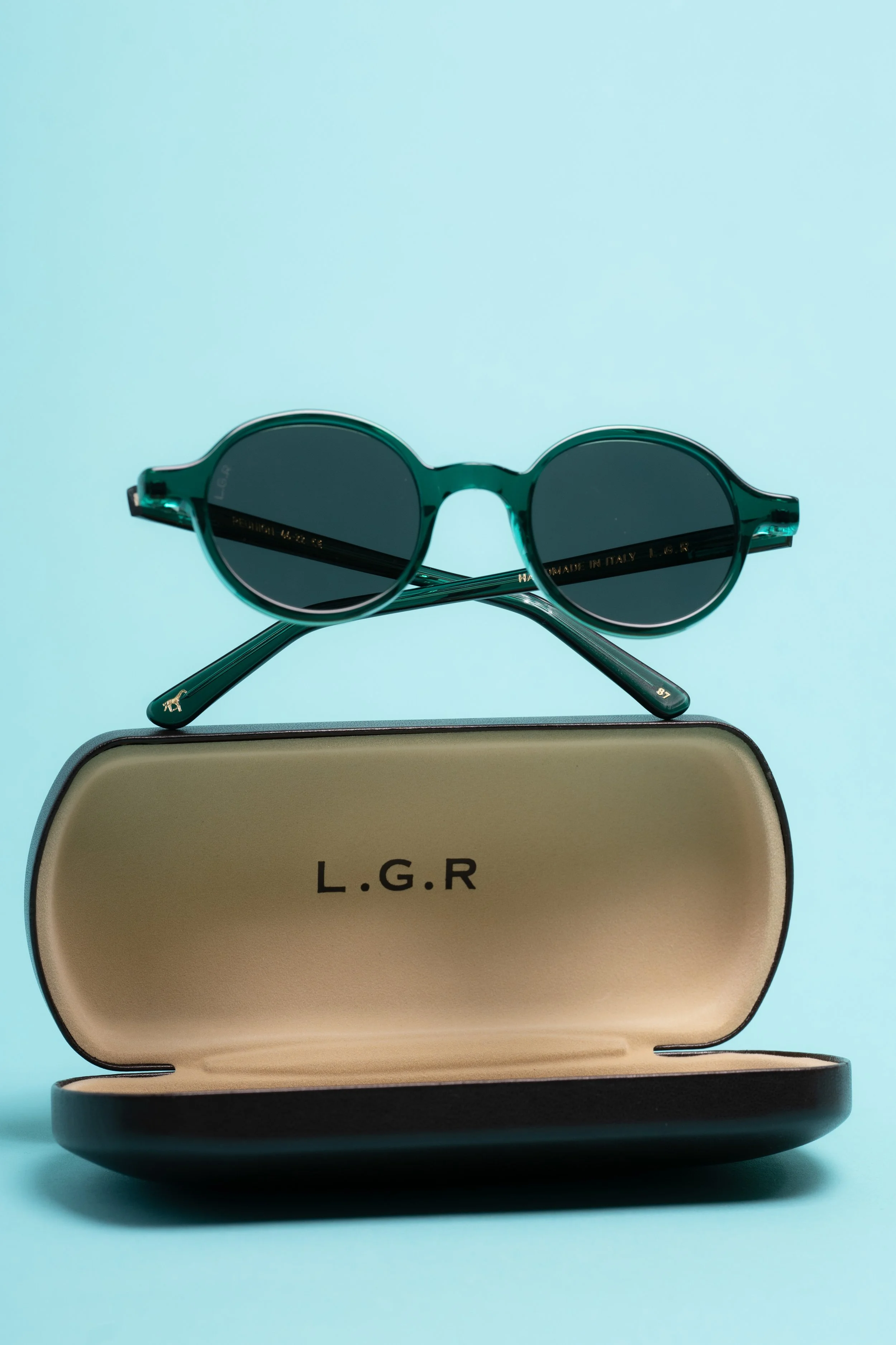 A pair of green sunglasses resting on a beige case with "L.G.R" written on it, set against a light blue background.