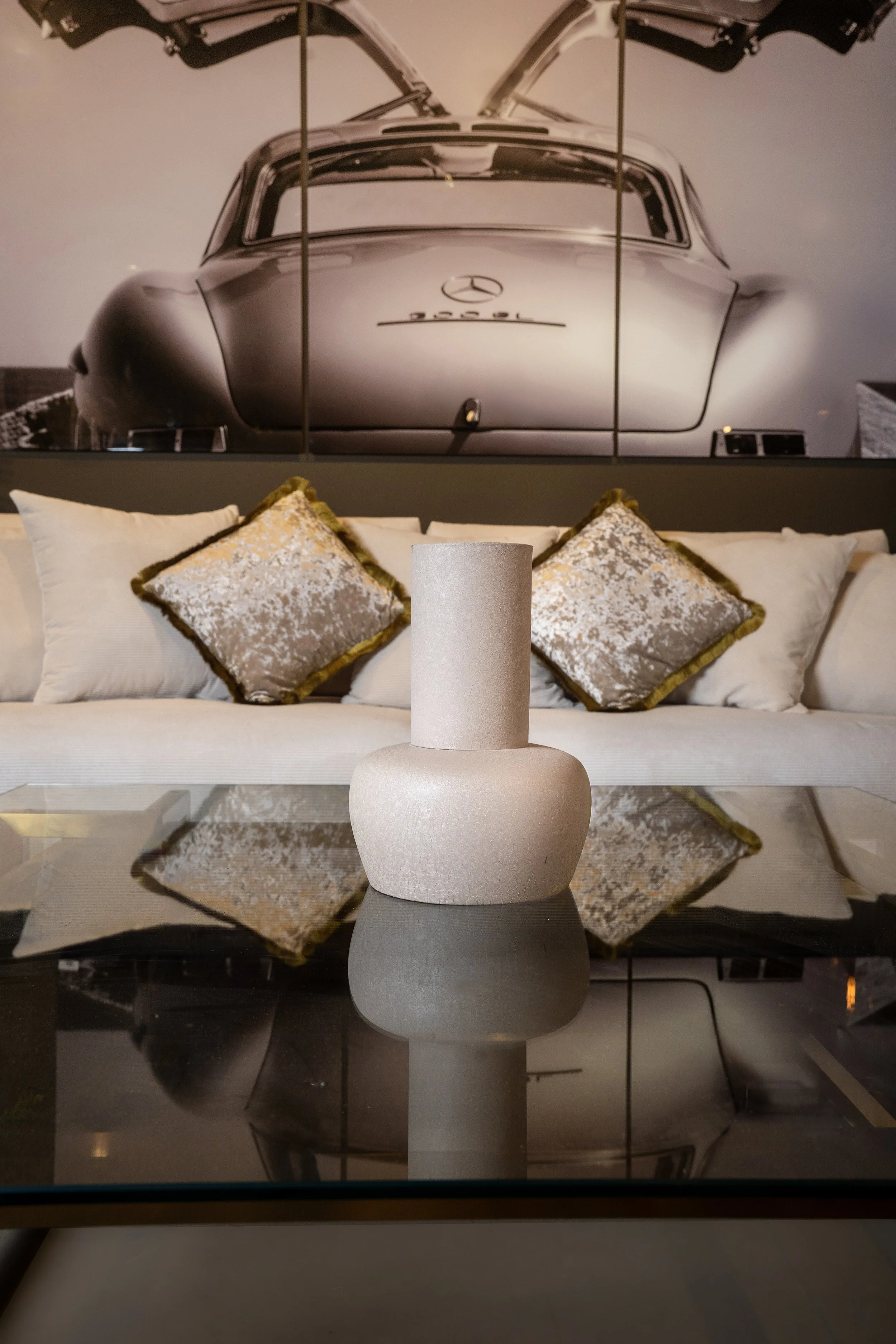 Modern living room with a glass coffee table, white textured vase, white velvet sofa, and decorative pillows, with a black-and-white vintage car wall art in the background.