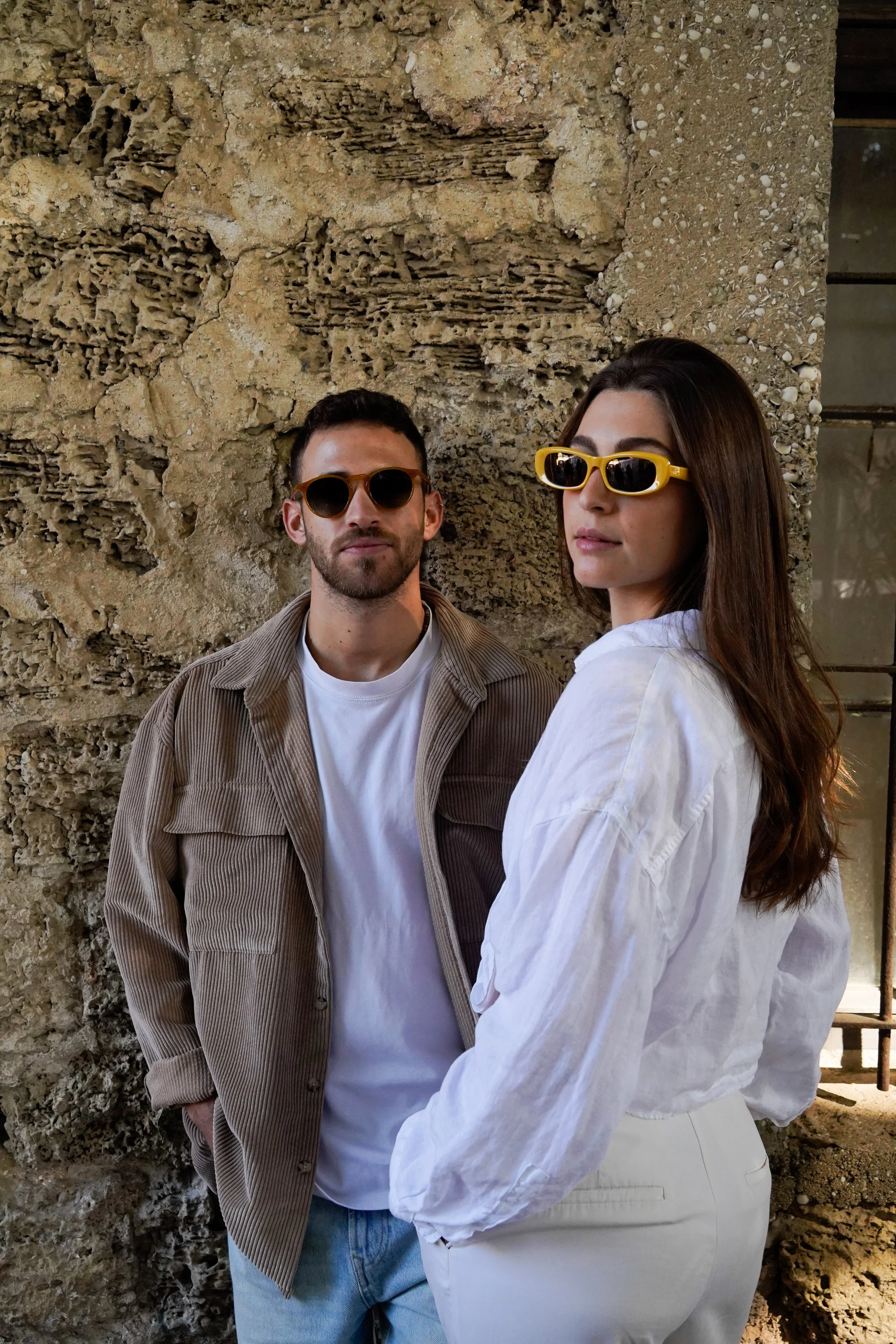 A man and woman standing in front of a textured stone wall, both wearing sunglasses, with the woman in a white blouse and the man in a brown jacket and white shirt.