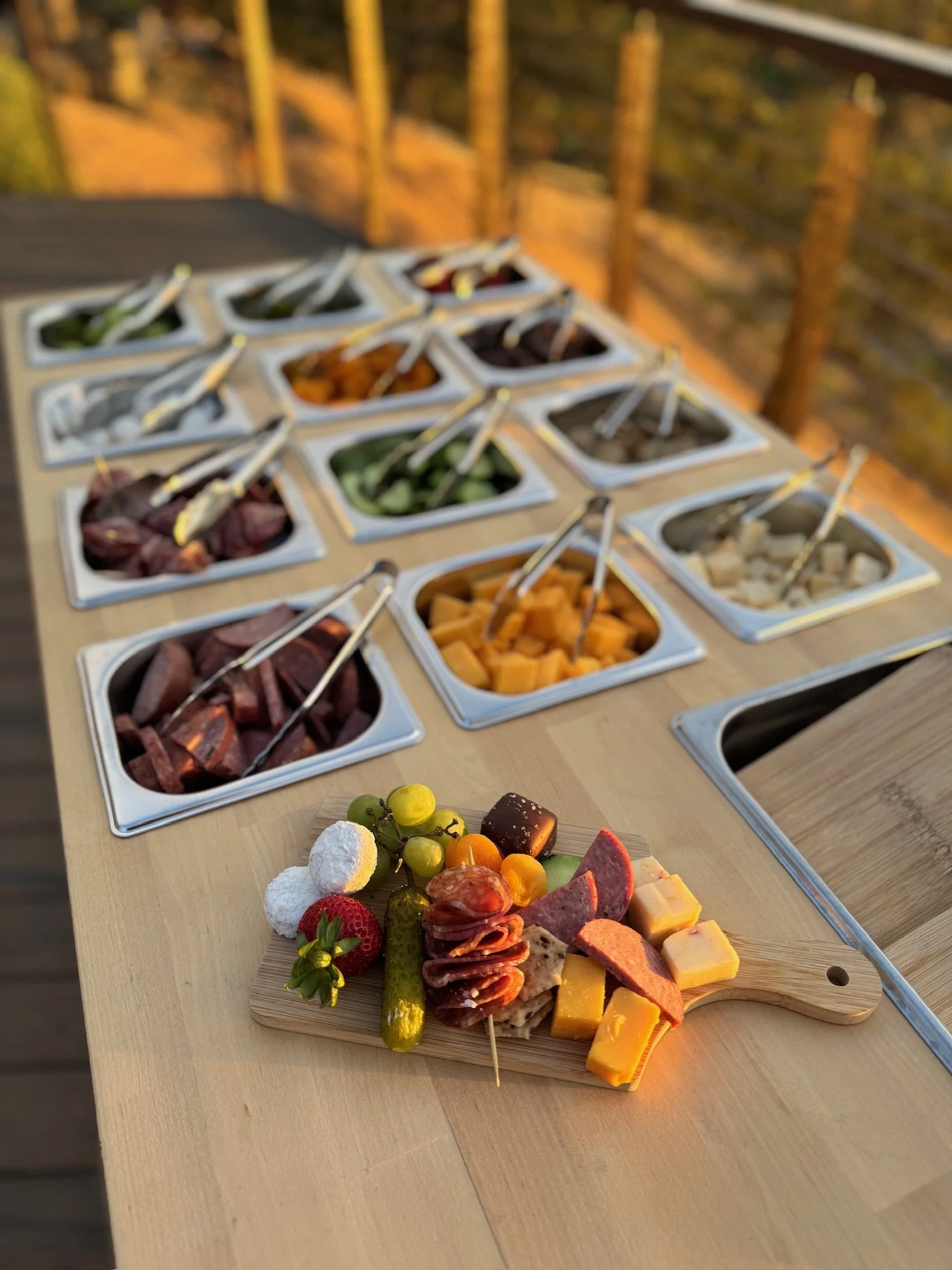 Charcuterie board with various cheese, meats, and fruits, set on a wooden table outside, with a background of a wooden deck and railing.