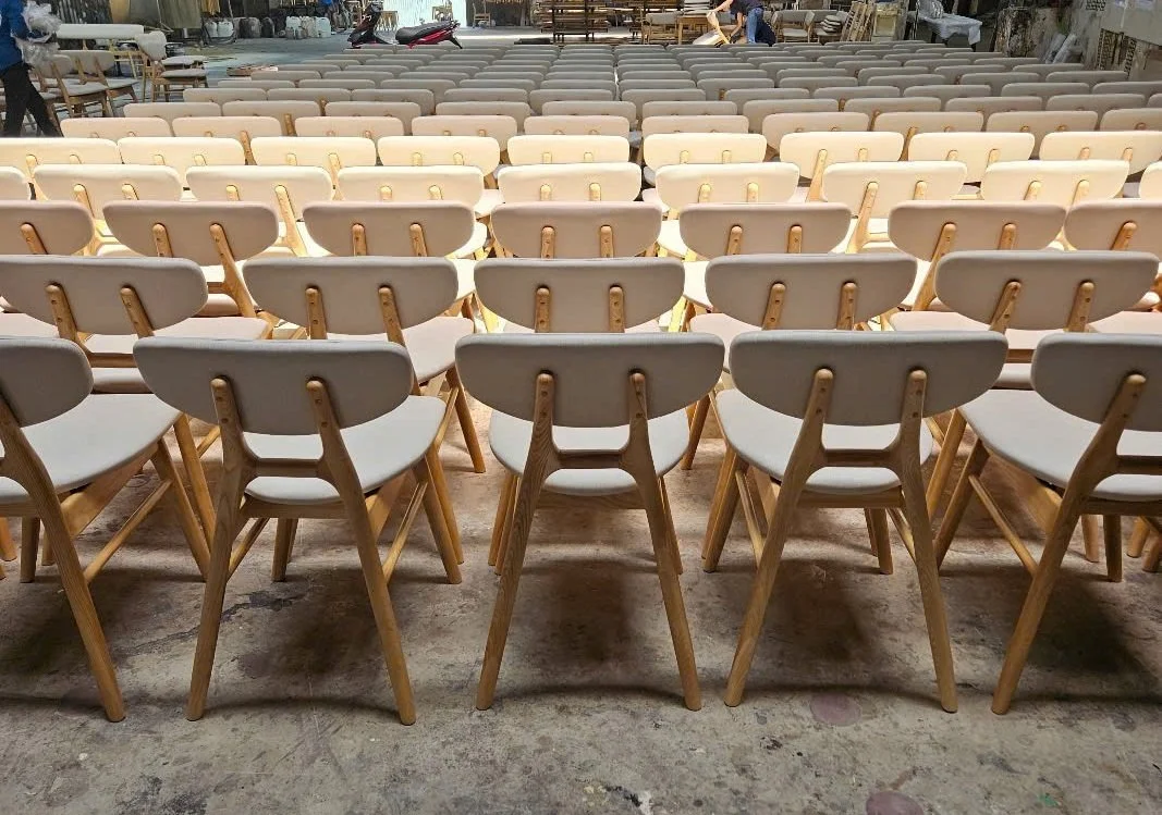 Multiple rows of beige chairs with wooden legs arranged in an orderly fashion, likely for an event or gathering, in a warehouse or storage facility with various furniture in the background.