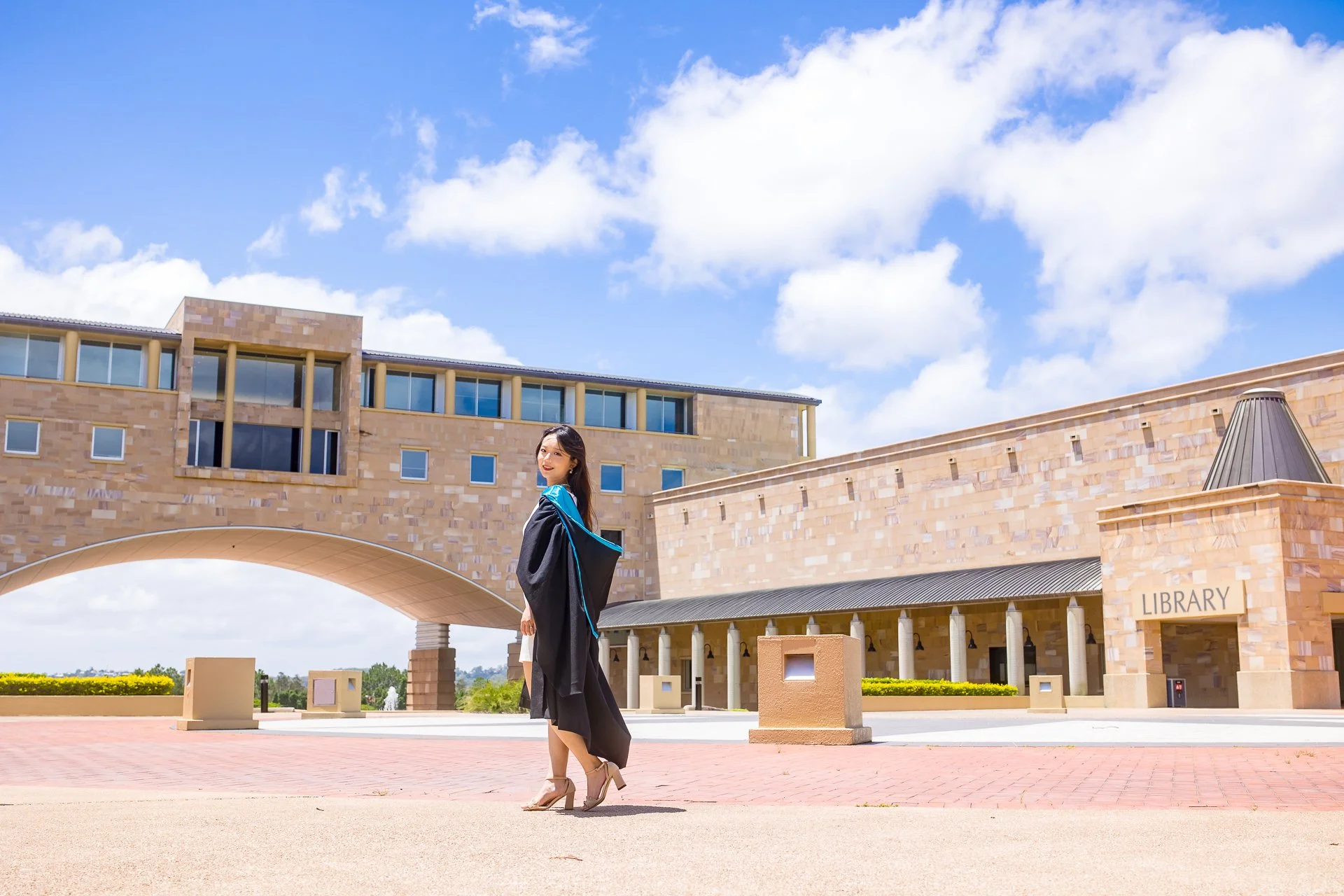 A young woman in a graduation gown standing outside a university library building on a sunny day.