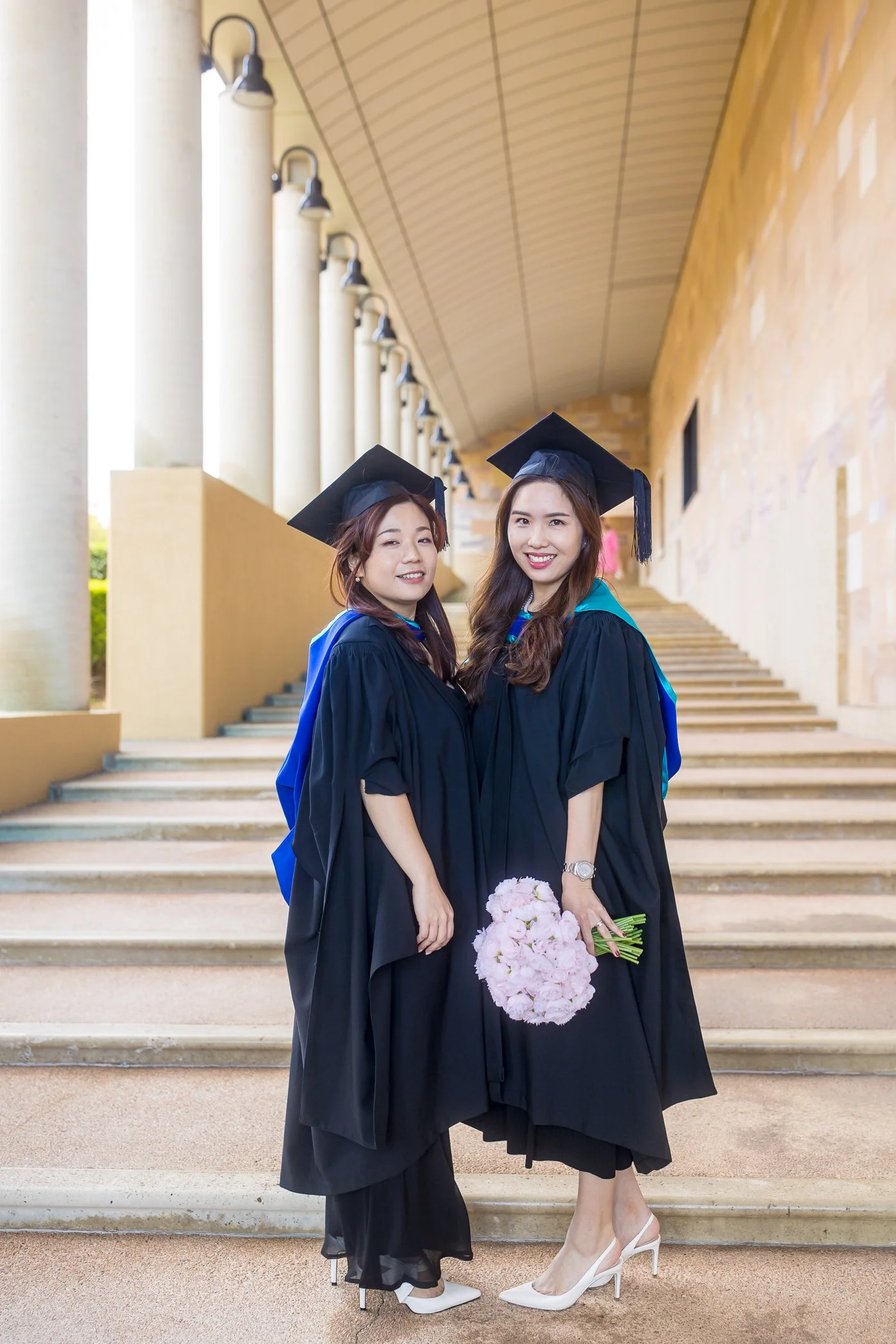 Two women in graduation caps and gowns standing on steps outside, smiling, one holding a bouquet of pink flowers.