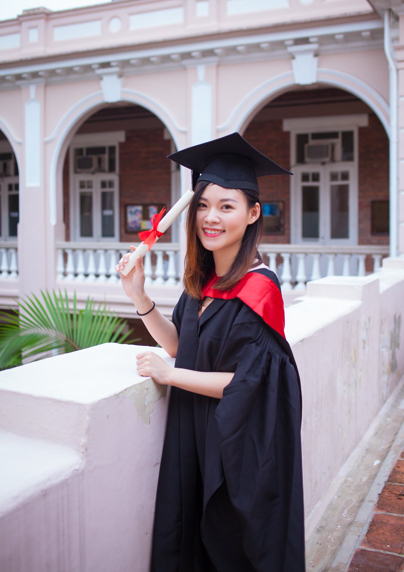 A young woman in graduation cap and gown smiling, holding a rolled diploma tied with a red ribbon, posing outdoors in front of a building with arches and windows.