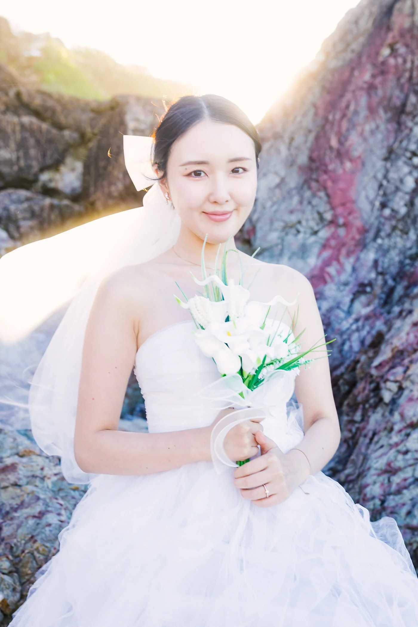 A woman in a white wedding dress holding a bouquet of white flowers, standing outdoors near rocks with sunlight in the background.