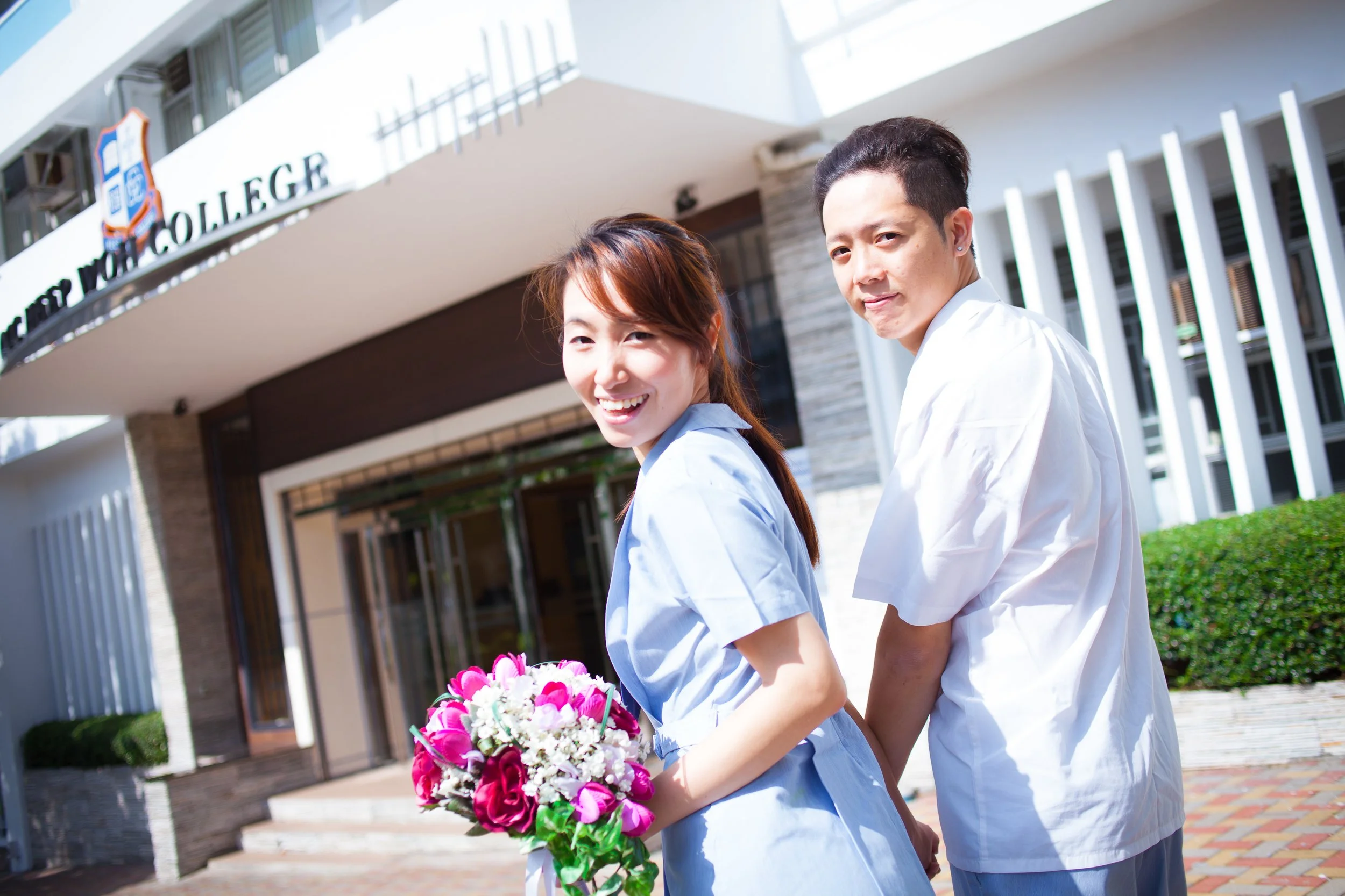 A couple holding hands and smiling outside a modern building that reads 'College of' with the rest of the sign cut off, one woman holding a bouquet of pink and white flowers, both dressed in light-colored casual clothing on a sunny day.