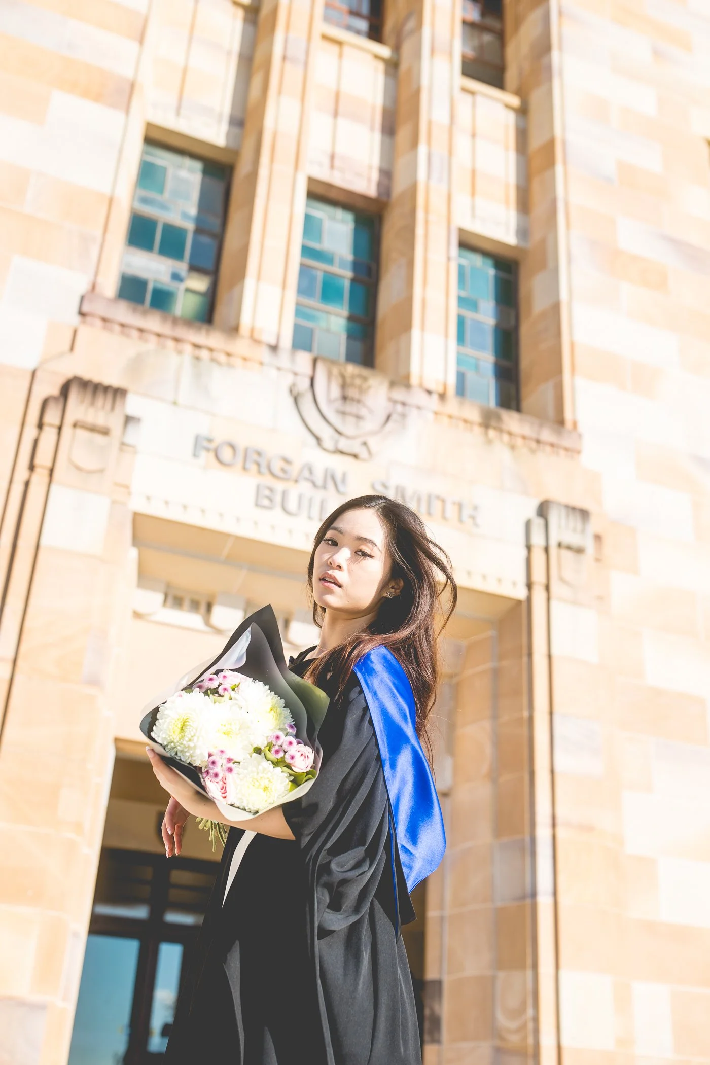 A young woman in a graduation gown holding a bouquet of flowers outside a university building