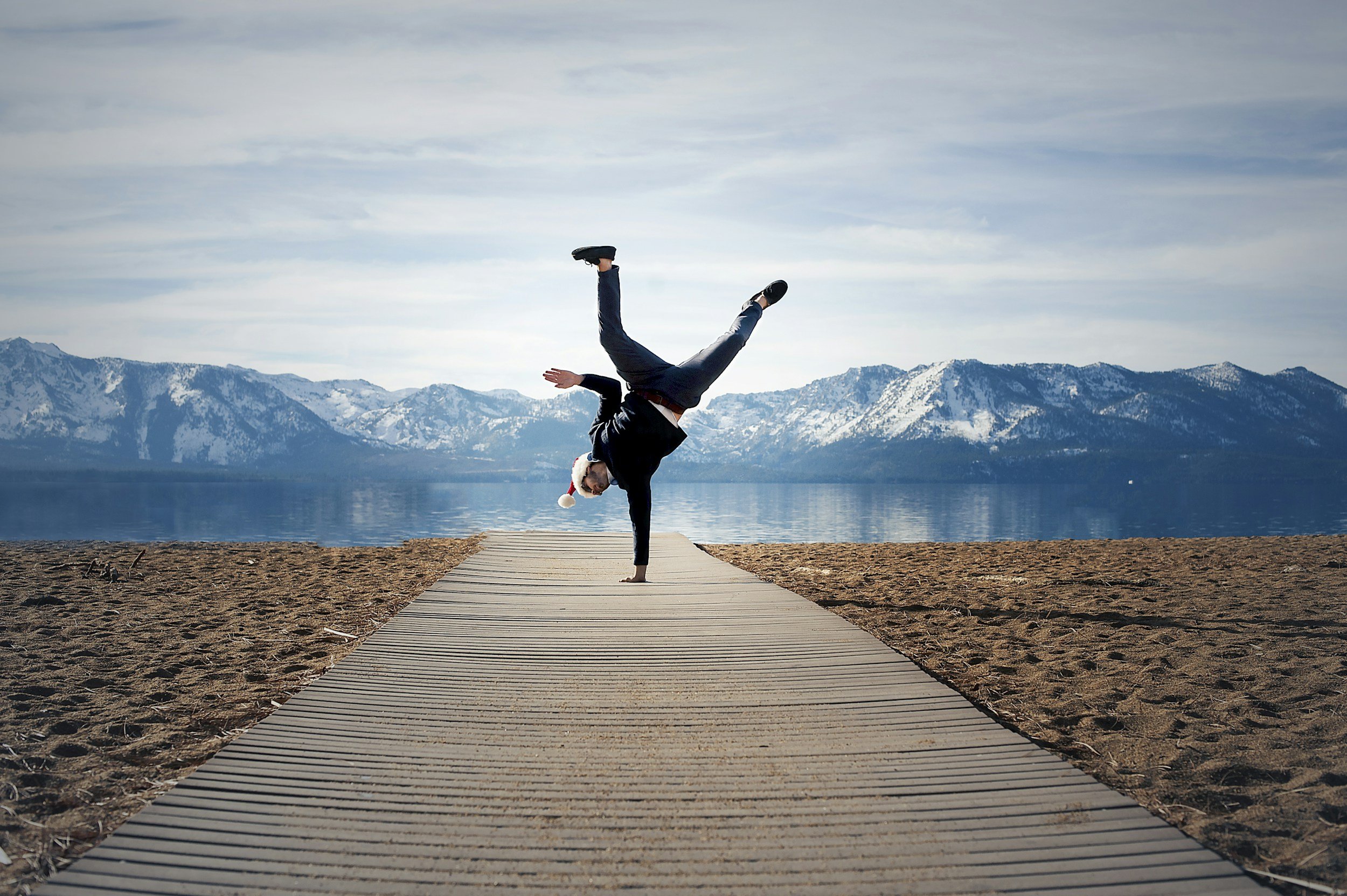 A person doing a handstand on a wooden pathway at the beach, wearing a Santa hat, with snow-capped mountains and a lake in the background.