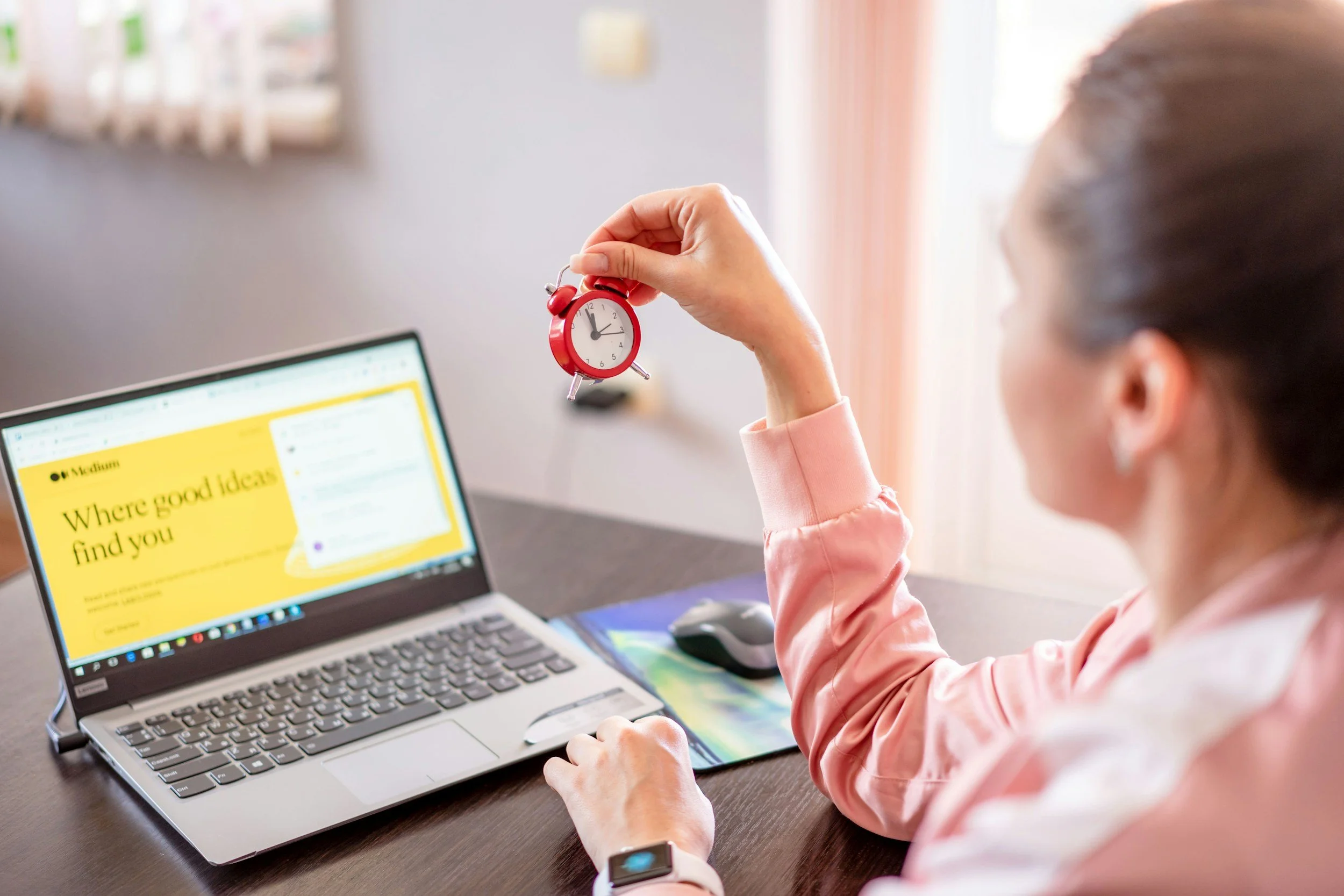 Woman sitting at a desk holding a small red alarm clock with a white face and black hands in front of a laptop displaying a yellow webpage.