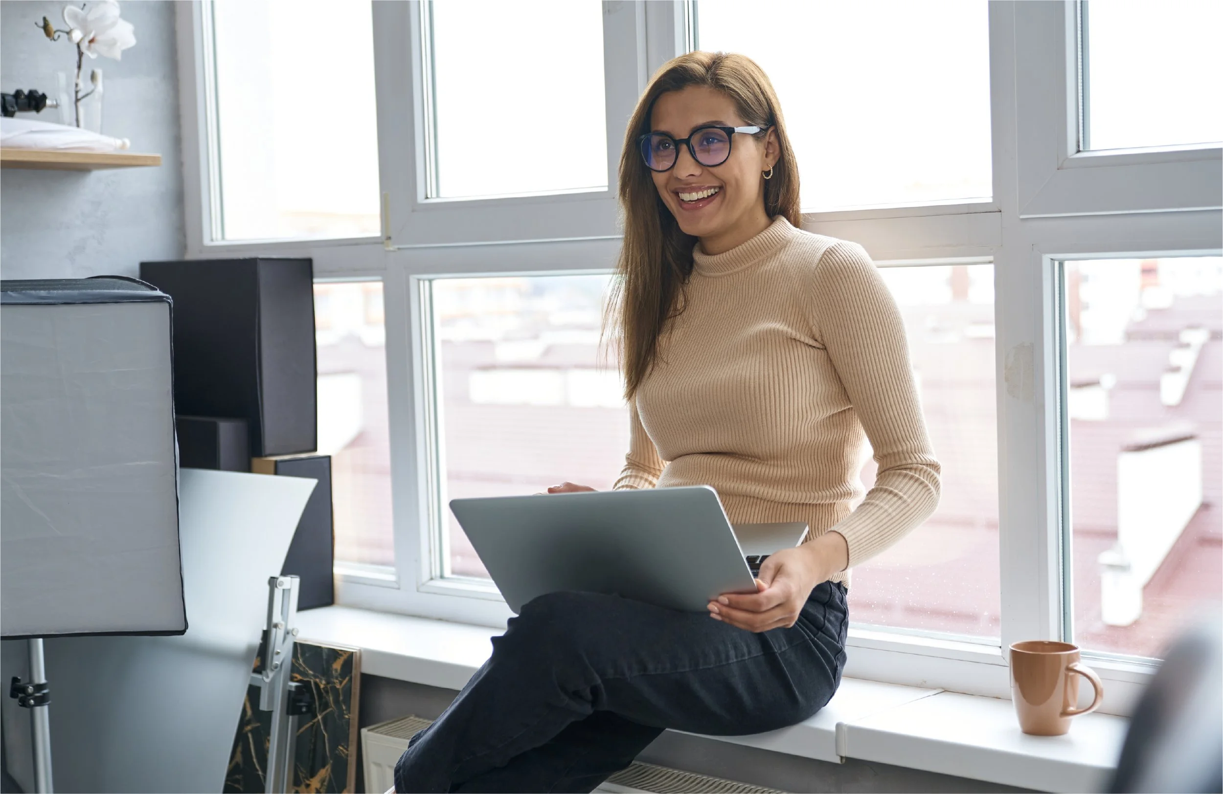A woman with glasses and a beige sweater sitting on a windowsill while holding a laptop and smiling in a well-lit room.