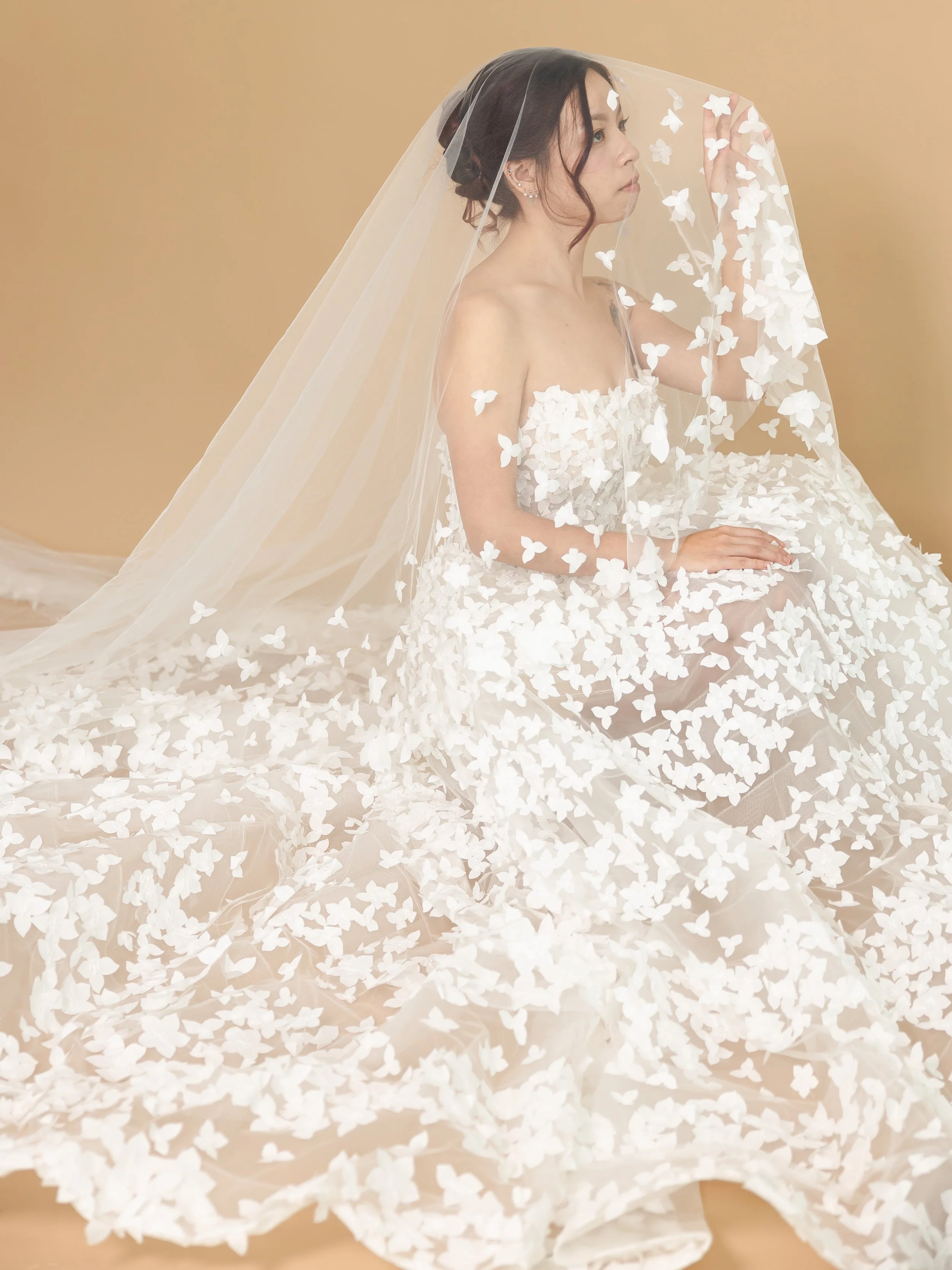 A woman dressed in a wedding gown with butterfly appliqués, sitting on the floor beneath a large veil, in front of a beige background.