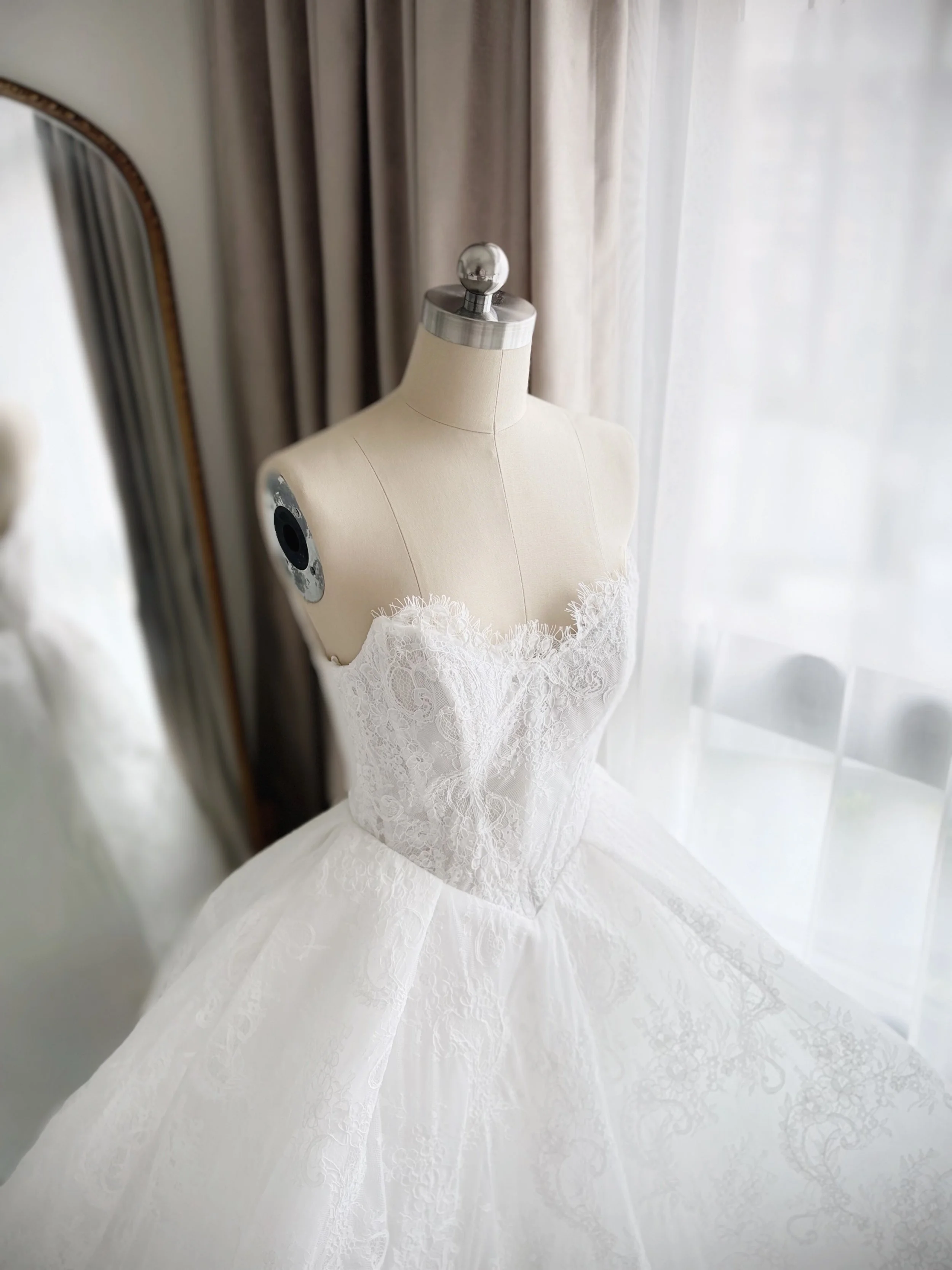 Wedding dress on a mannequin near a mirror and window with curtains.