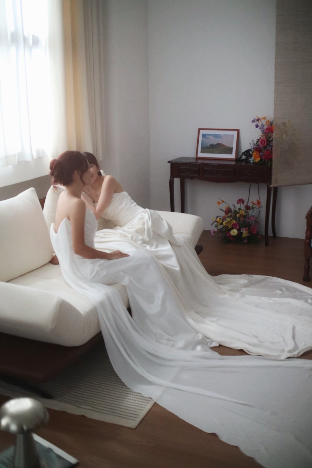 Two women in white wedding dresses sitting on a couch in a room with a window, a wooden table with a framed picture, flowers, and a black telephone.
