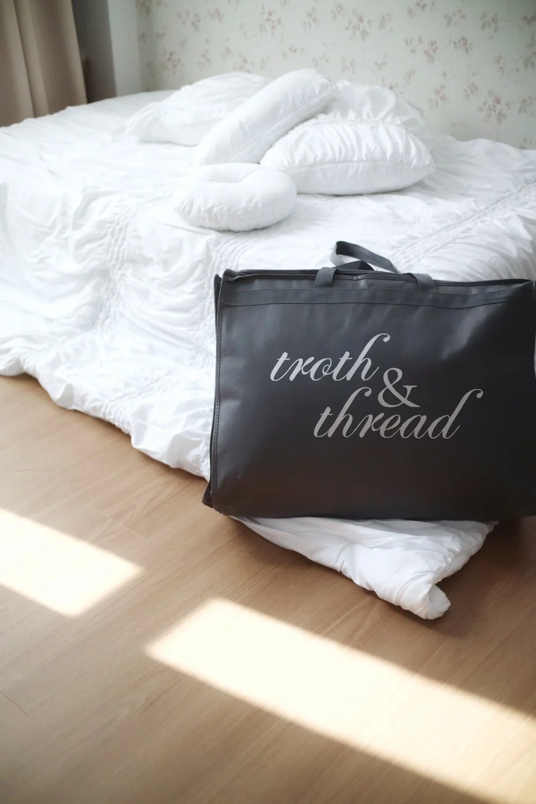 A black garment bag with the words "troth & thread" on it, resting against a bed with white bedding and pillows in a room with floral wallpaper and beige curtains.