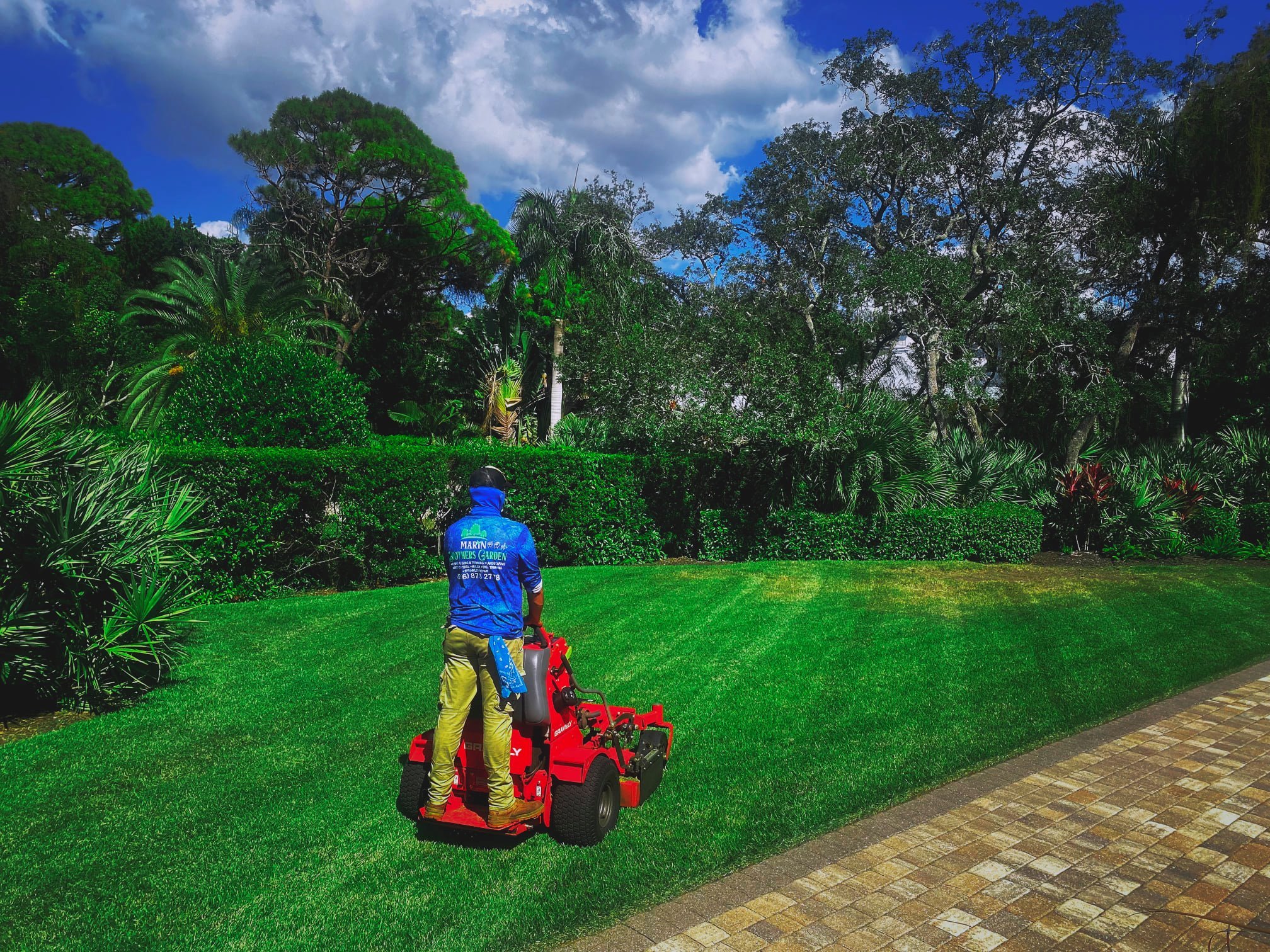 A person wearing a blue long sleeve shirt, khaki pants, and a black cap is operating a red lawn mower on a green lawn bordered by a stone pathway, with lush trees and bushes under a partly cloudy sky.