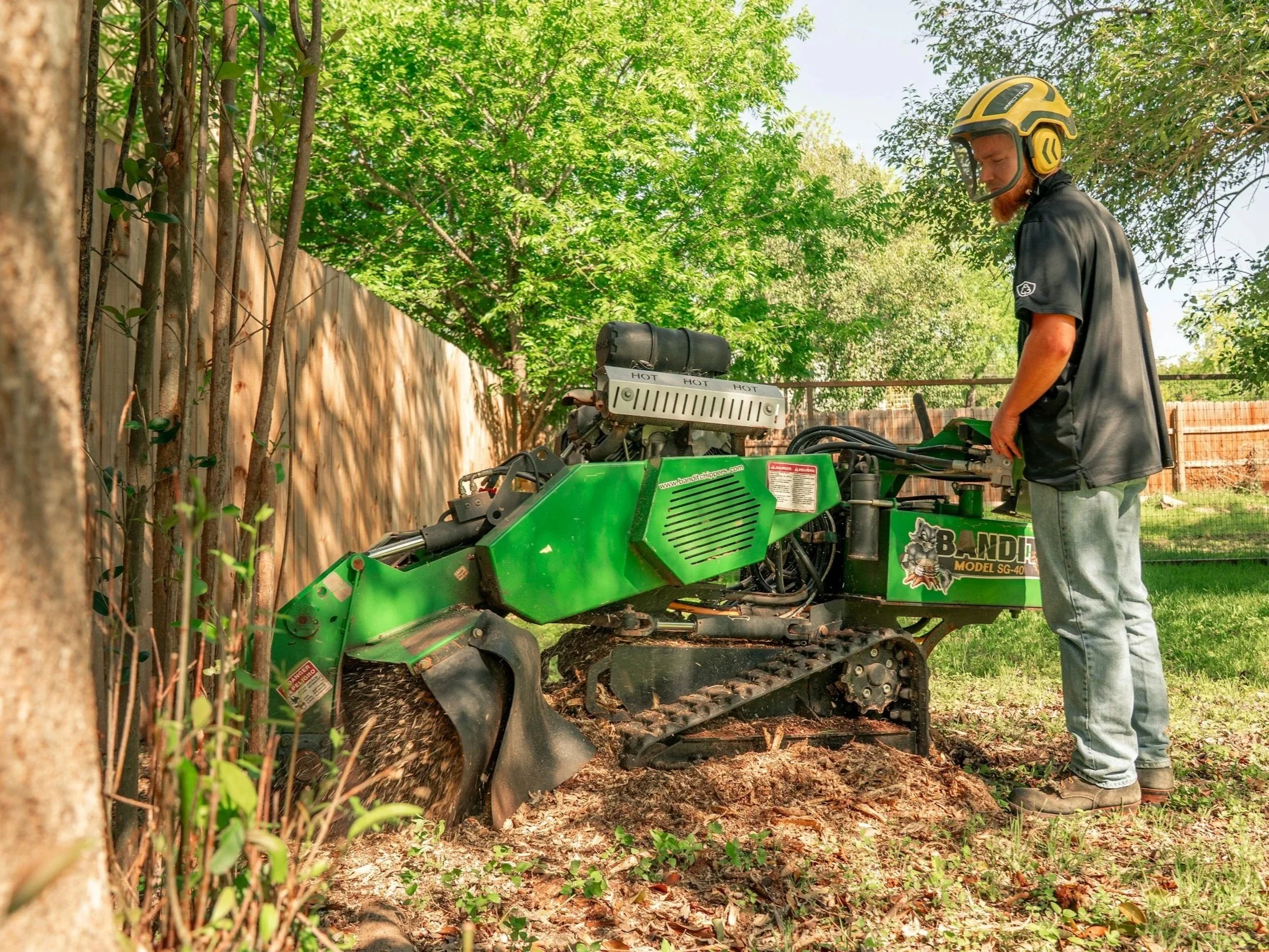 Man operating a green wood chipper in a backyard with trees and a wooden fence.