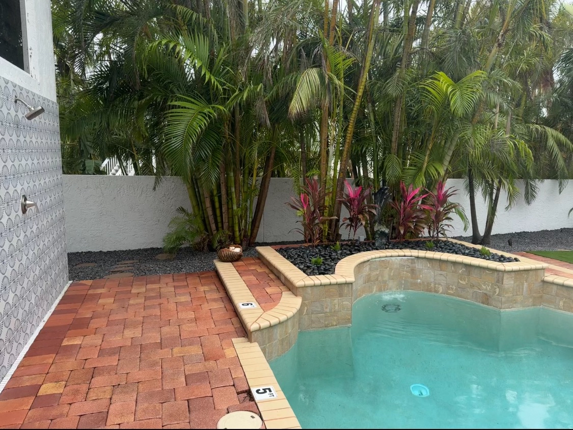 A backyard pool area with a brick deck, surrounded by tropical plants, including palm trees and pink-leafed plants, with a white wall and outdoor shower on the left.
