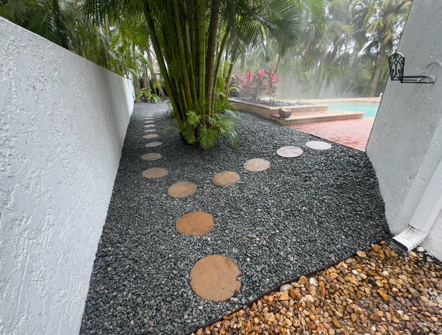 A backyard pathway with circular stepping stones passing through black gravel, beside white textured walls, leading to a pool area with red brick pavement, greenery, and mist around the pool.