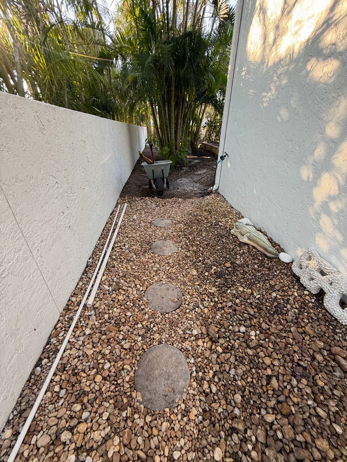 A narrow outdoor pathway with pebble stones and round stepping stones, flanked by white stucco walls, with a wheelbarrow and gardening tools at the end, surrounded by green plants and trees.