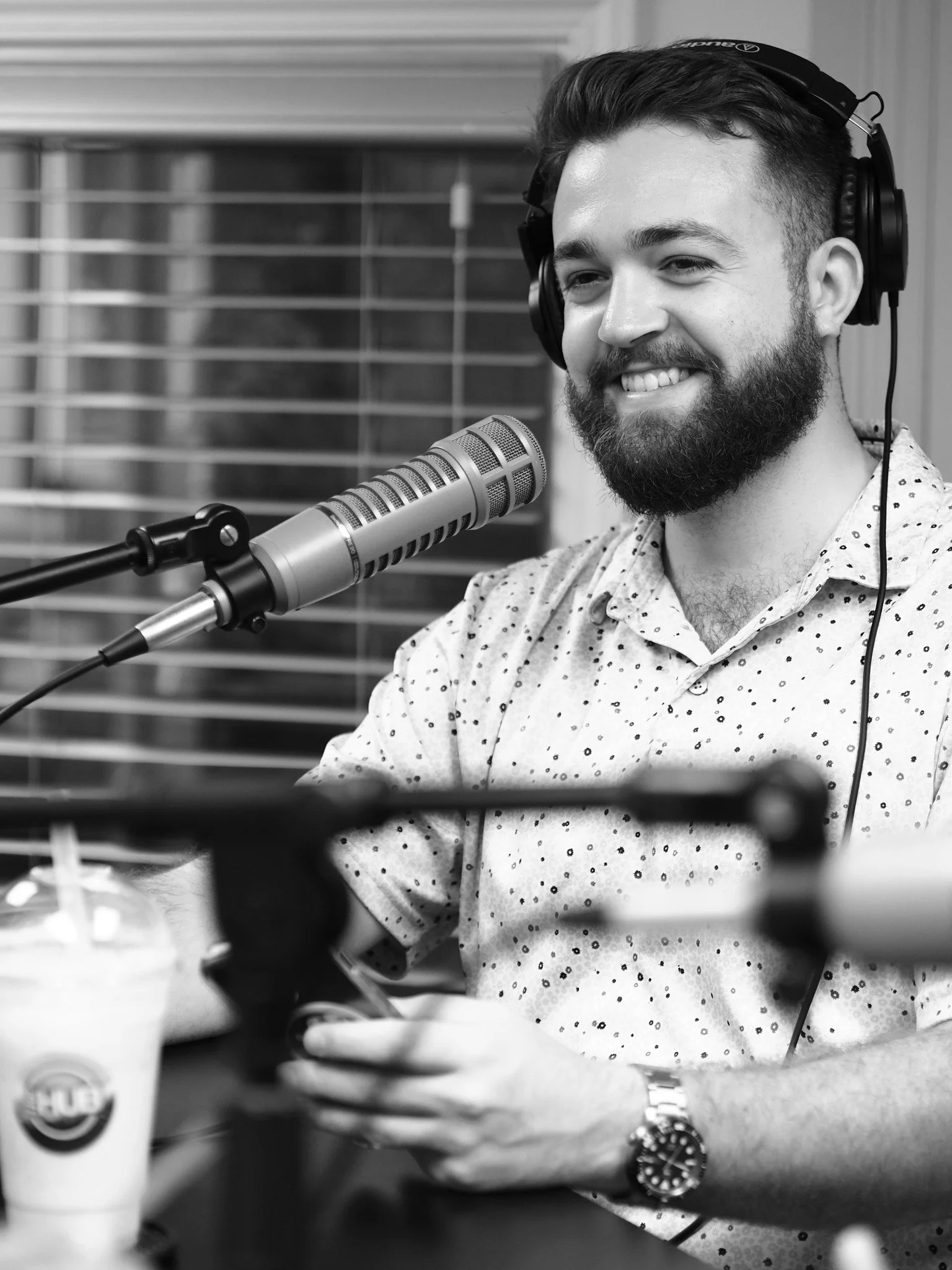 A man with a beard and short hair, wearing headphones, smiling, sitting at a table with a microphone and a drink, in a recording studio.