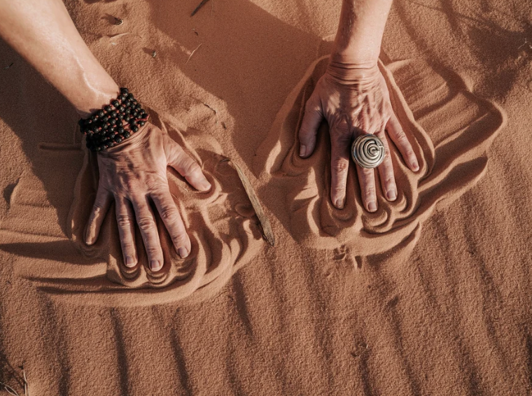 Two hands pressing into the sand, creating impressions, with one hand wearing a shell ring and the other wearing beaded bracelets.