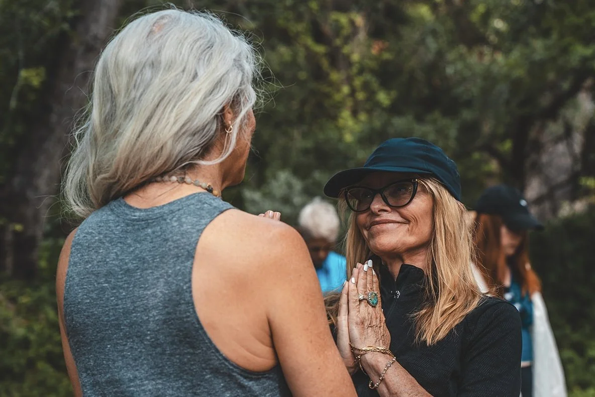 Two women standing close, one with gray hair and the other with long blonde hair, wearing glasses and a black hat, engaging in a peaceful moment outdoors.