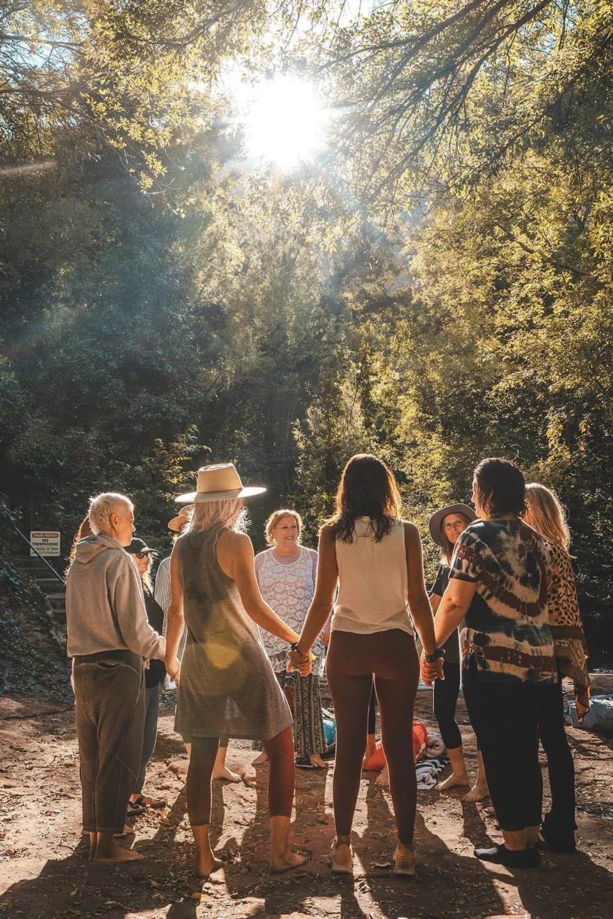 A group of people holding hands in a circle outdoors, with sunlight shining through trees overhead, creating a warm and peaceful atmosphere.