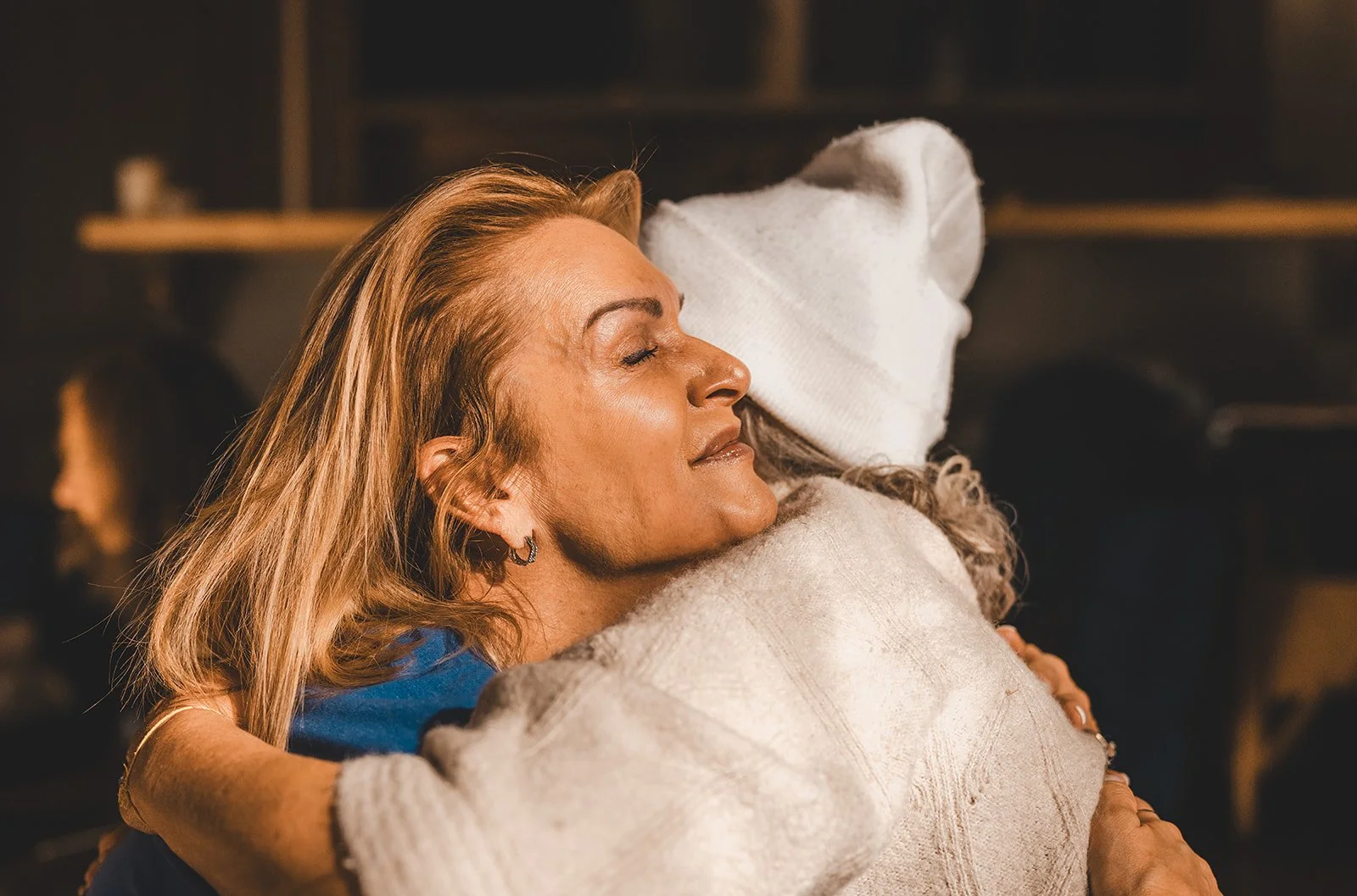 Two women hugging warmly, one with blonde hair and the other with curly hair wearing a white beanie, in a cozy indoor setting.