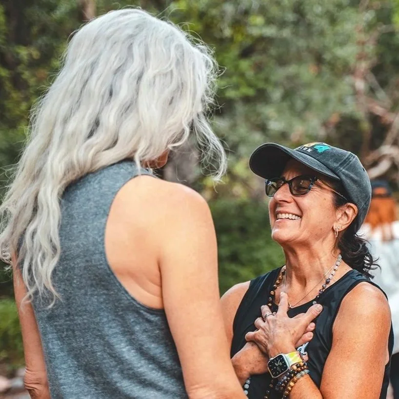 Two women outdoors, one with long gray hair and the other with short dark hair, wearing glasses and a baseball cap, happily talking and smiling.
