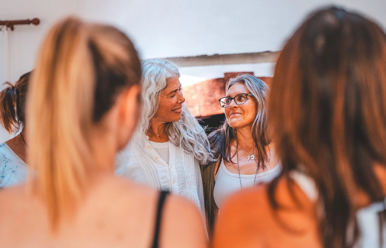 Two women are smiling and looking at each other, surrounded by other women, in an indoor setting with a window in the background.