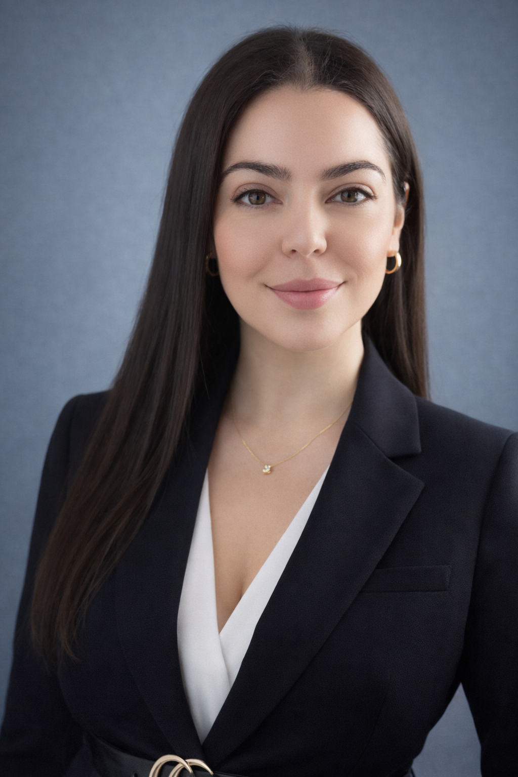 A young woman with long dark hair, wearing a black blazer and a white top, smiling, with gold hoop earrings and a delicate gold necklace, against a plain grey background.
