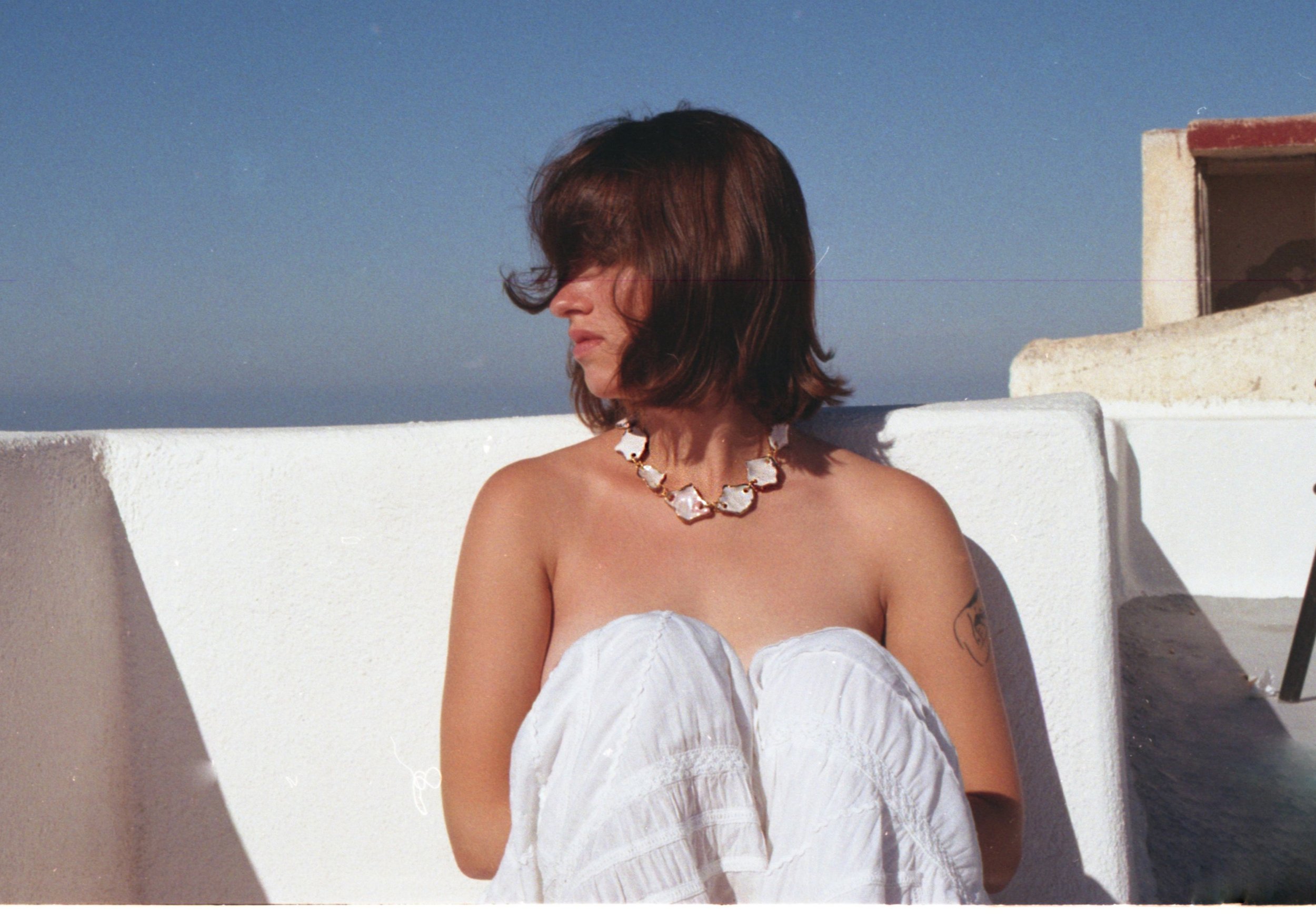 Femme aux cheveux bruns portant un collier blanc, assise avec les genoux levés, face à un mur blanc sur fond de ciel bleu, dans un lieu ensoleillé.
