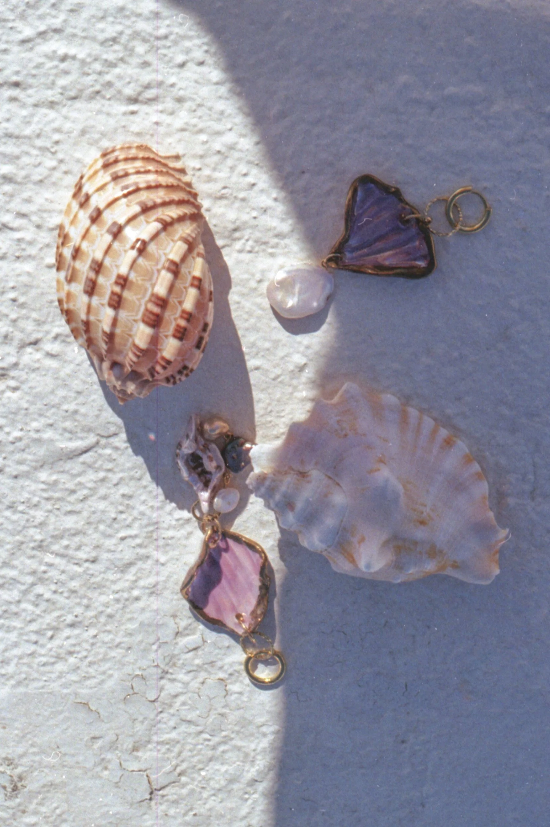 Des bijoux en forme de coquillages et de pierres pourpres et roses, disposés sur une surface blanche et grise avec un éclairage naturel.