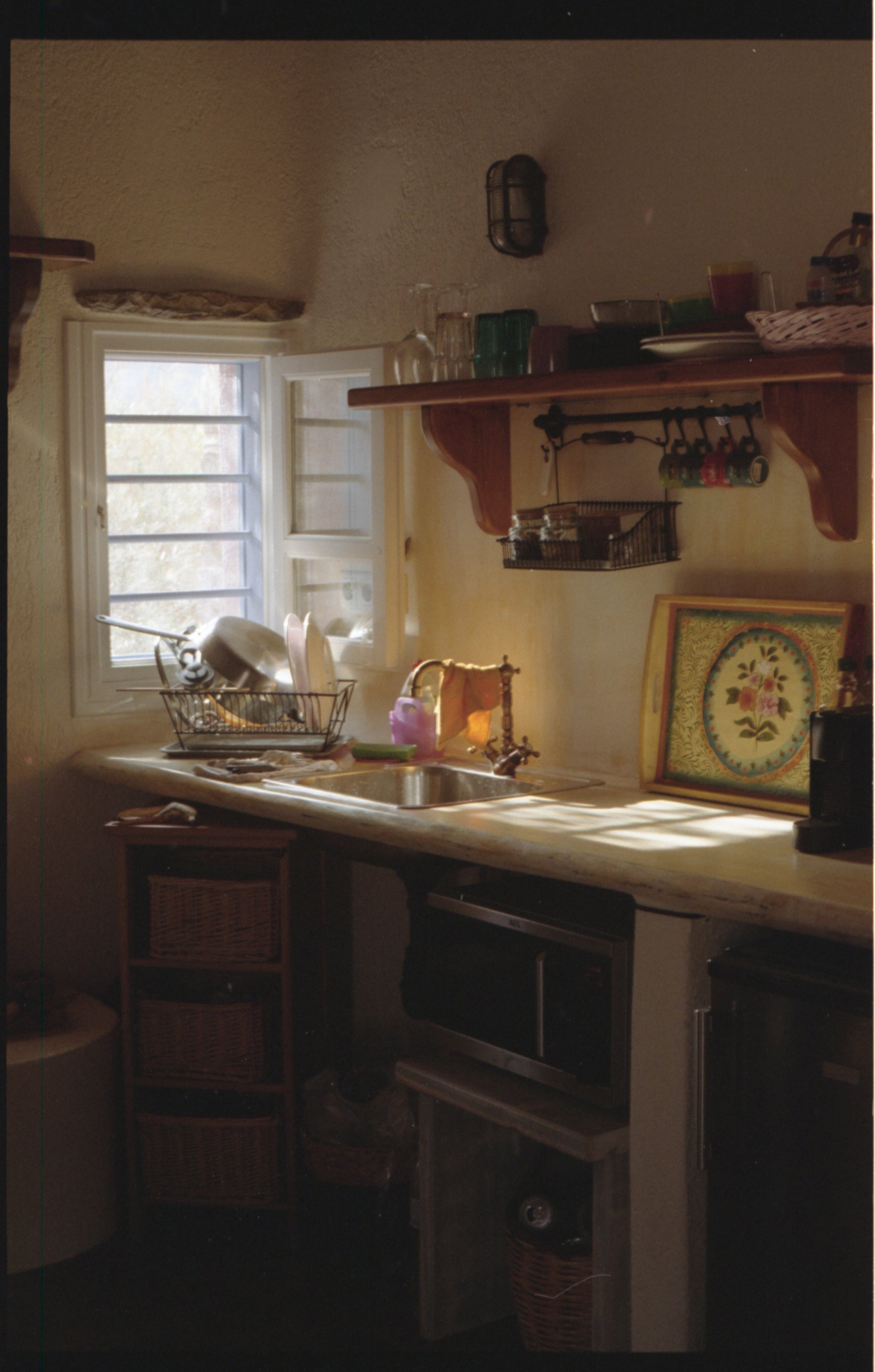 Une cuisine rustique avec une fenêtre, un évier en acier inoxydable, une étagère en bois avec divers objets, une caisse en bois avec 3 tiroirs, un plateau décoratif avec un motif floral, et des ustensiles de cuisine.