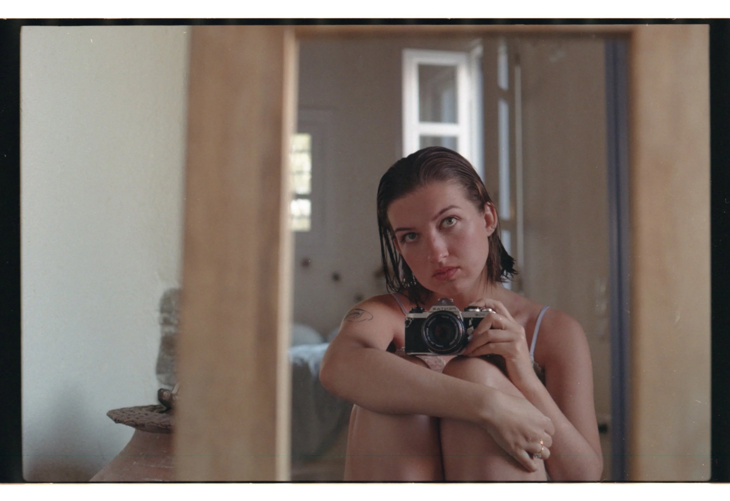 Une jeune femme aux cheveux bruns courts, portant un débardeur, assise devant un miroir, tenant un appareil photo, dans une pièce lumineuse, vue dans un miroir