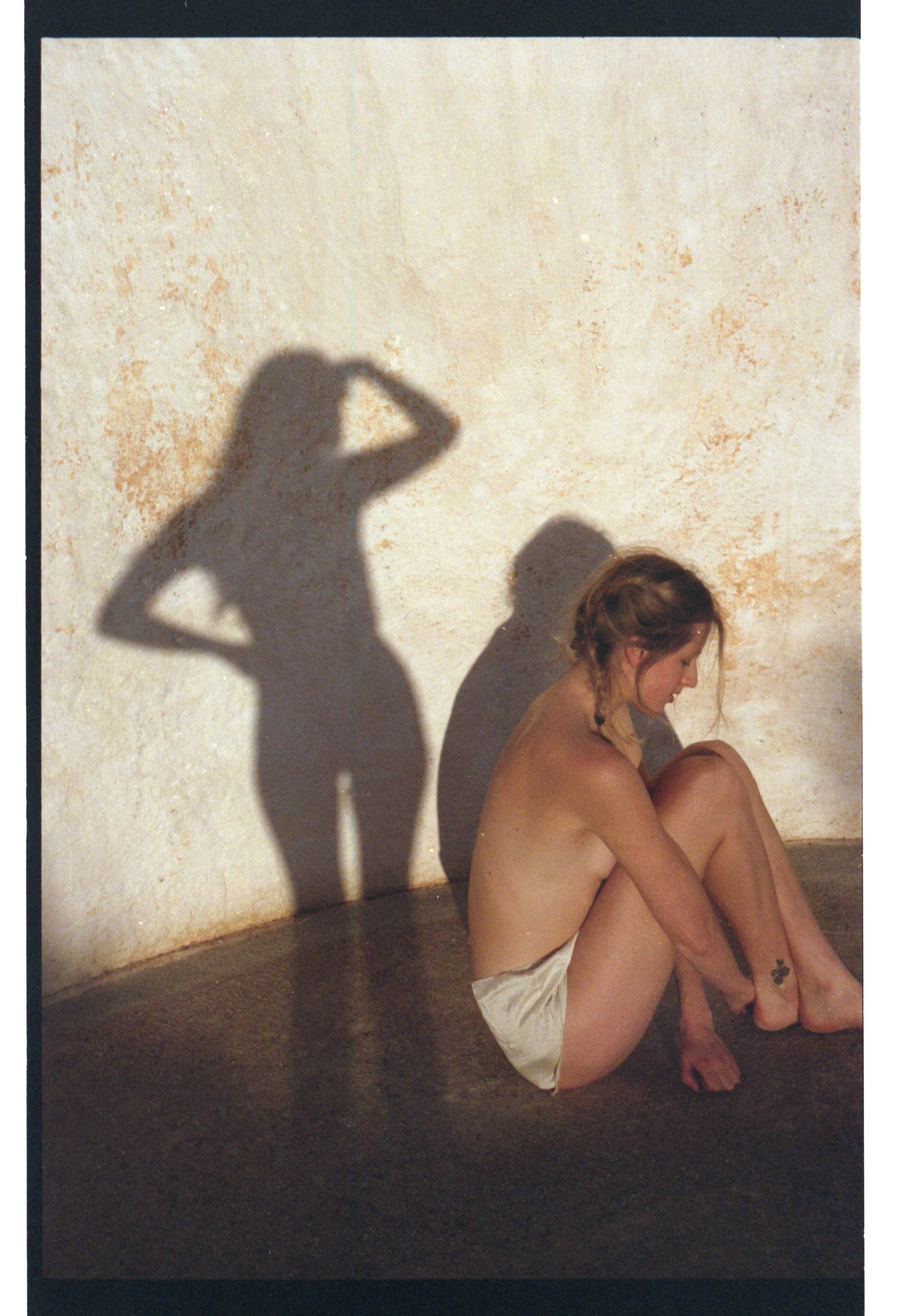Une femme nue assise contre un mur, regardant vers le sol, avec un tatouage de trèfle sur la cheville. La lumière crée une ombre de sa silhouette sur le mur derrière elle.